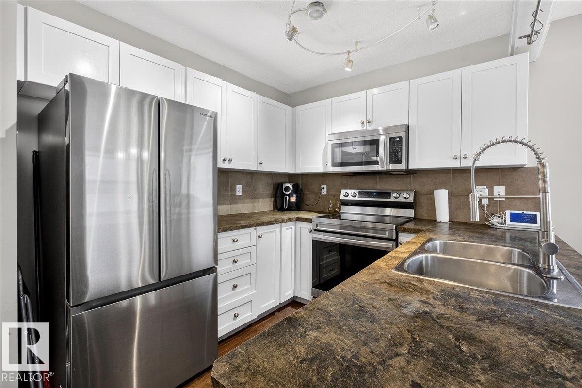 Kitchen featuring stainless steel appliances, white cabinetry, backsplash, and dark stone counters - 47 230 Edwards Drive, Edmonton, AB - Indoor Photo Showing Kitchen With Double Sink