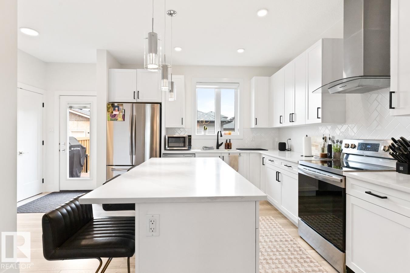 Modern kitchen featuring a large central island, white cabinetry with black hardware, stainless steel appliances, a herringbone tile backsplash, and light wood-finish flooring - 6023 180 Avenue, Edmonton, AB - Indoor Photo Showing Kitchen With Stainless Steel Kitchen With Upgraded Kitchen