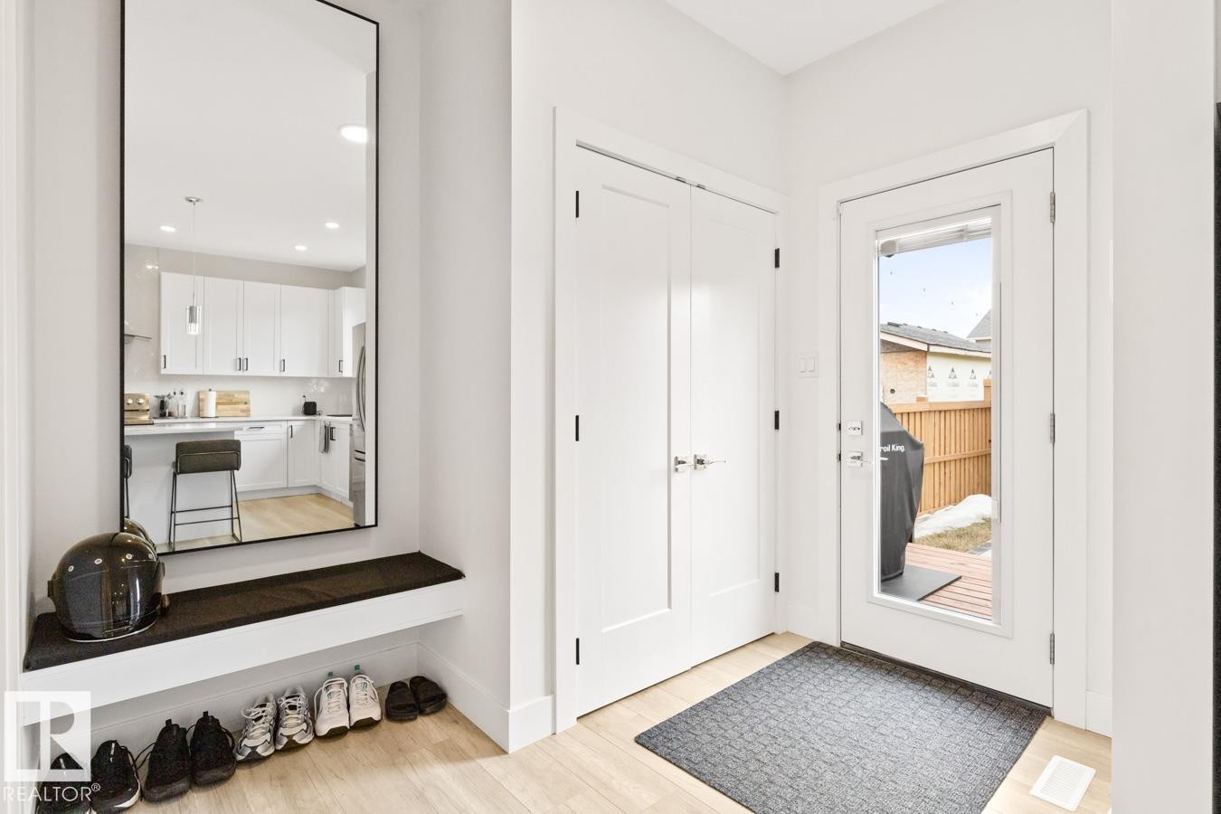 Entryway featuring light wood-finish flooring, a built-in storage bench with a dark cushion, a full-length mirror, and a coat closet with double doors - 6023 180 Avenue, Edmonton, AB - Indoor Photo Showing Other Room