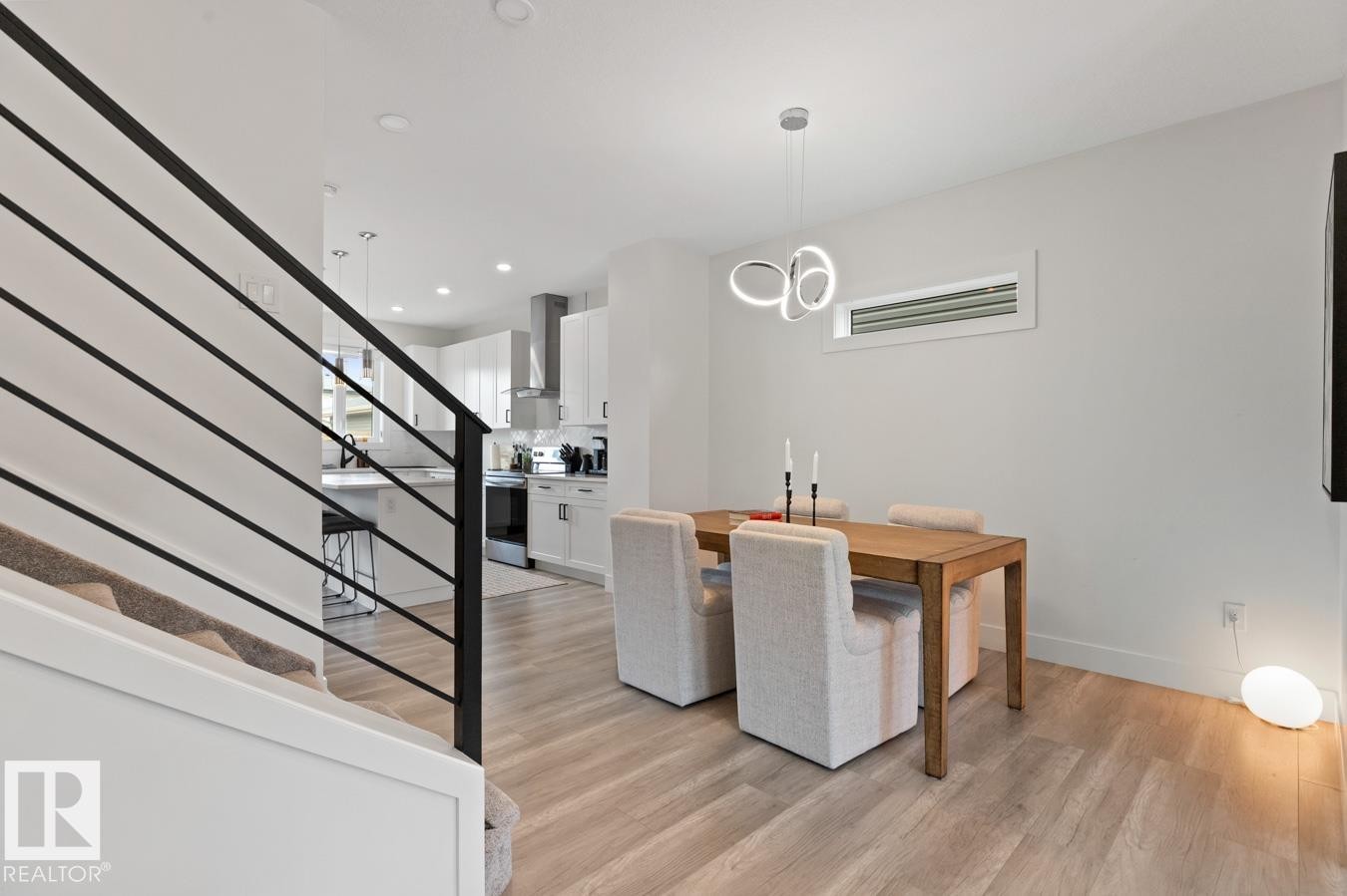 Open concept living space featuring wood-finish flooring, a contemporary chandelier, a horizontal window, and a sleek black metal staircase railing - 6023 180 Avenue, Edmonton, AB - Indoor Photo Showing Other Room