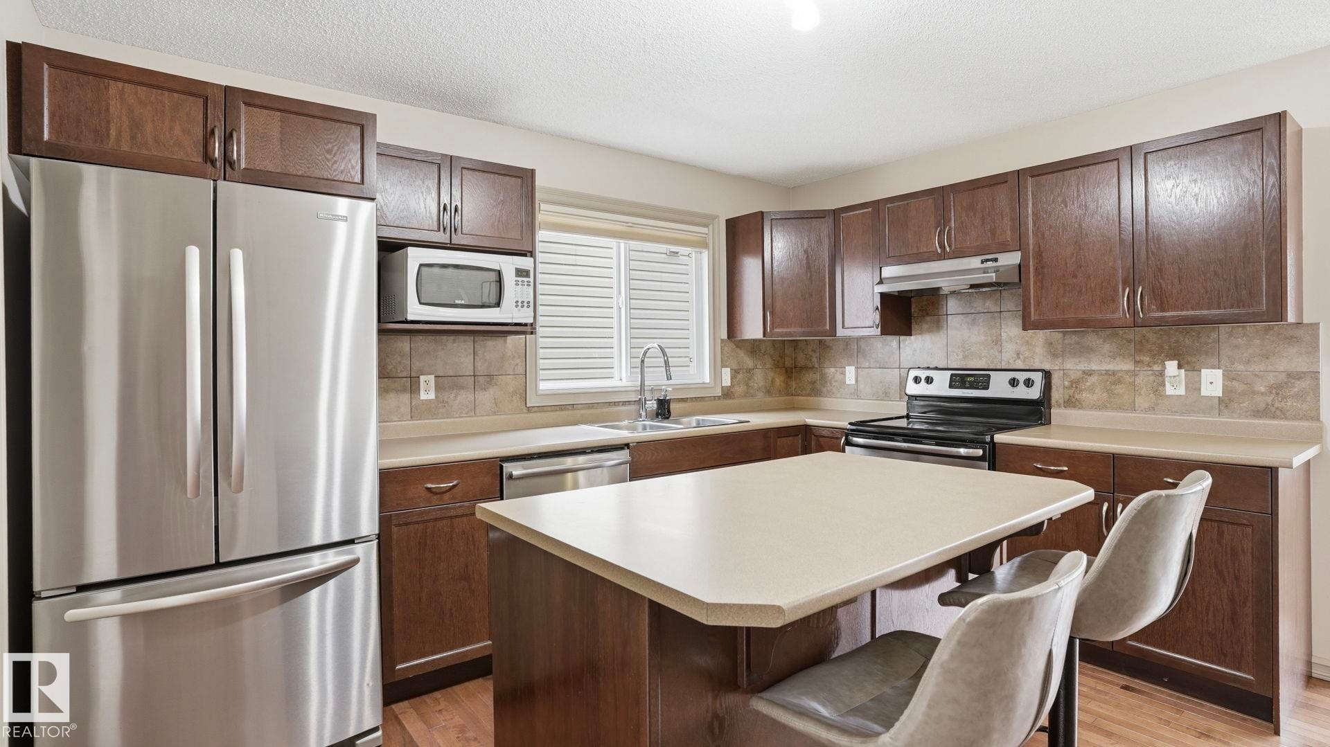 Kitchen featuring dark wood-finish cabinetry, stainless steel appliances, an island with countertop, and tile backsplash - 8210 180 Avenue Nw, Edmonton, AB - Indoor Photo Showing Kitchen With Double Sink