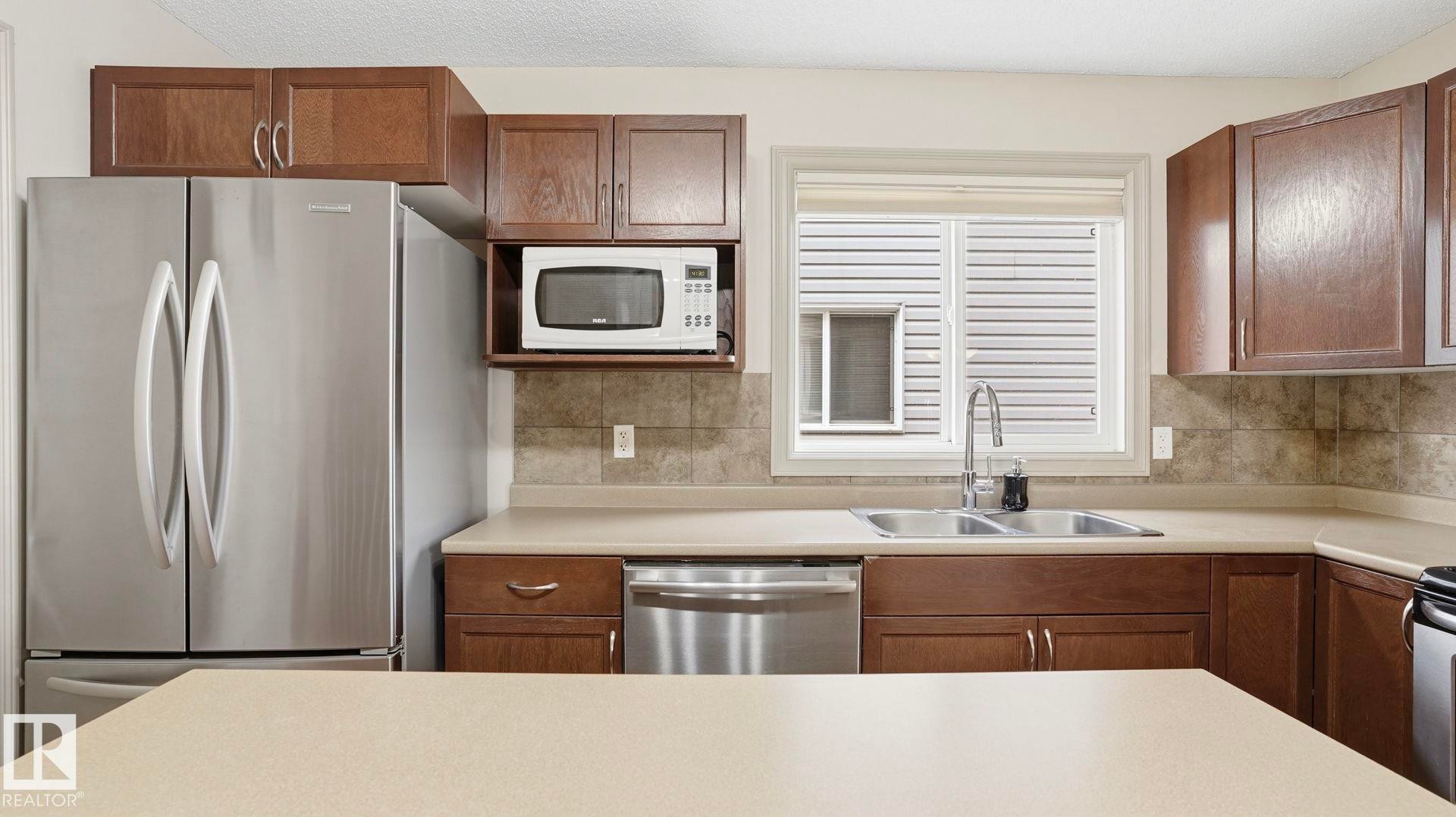 Kitchen featuring abundant cabinetry, stainless steel appliances, dual basin sink with gooseneck faucet, light-toned countertops, and a tiled backsplash - 8210 180 Avenue Nw, Edmonton, AB - Indoor Photo Showing Kitchen With Double Sink