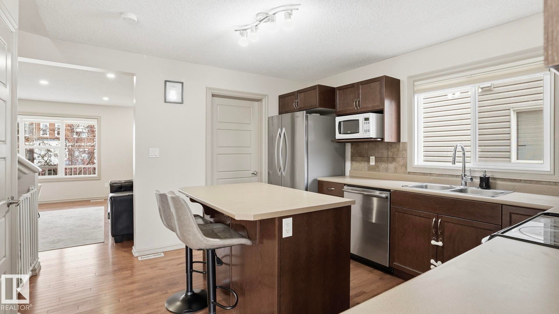 Kitchen featuring wood-finish flooring, dark wood cabinetry, light-toned countertops, and stainless steel appliances - 8210 180 Avenue Nw, Edmonton, AB - Indoor Photo Showing Kitchen With Double Sink