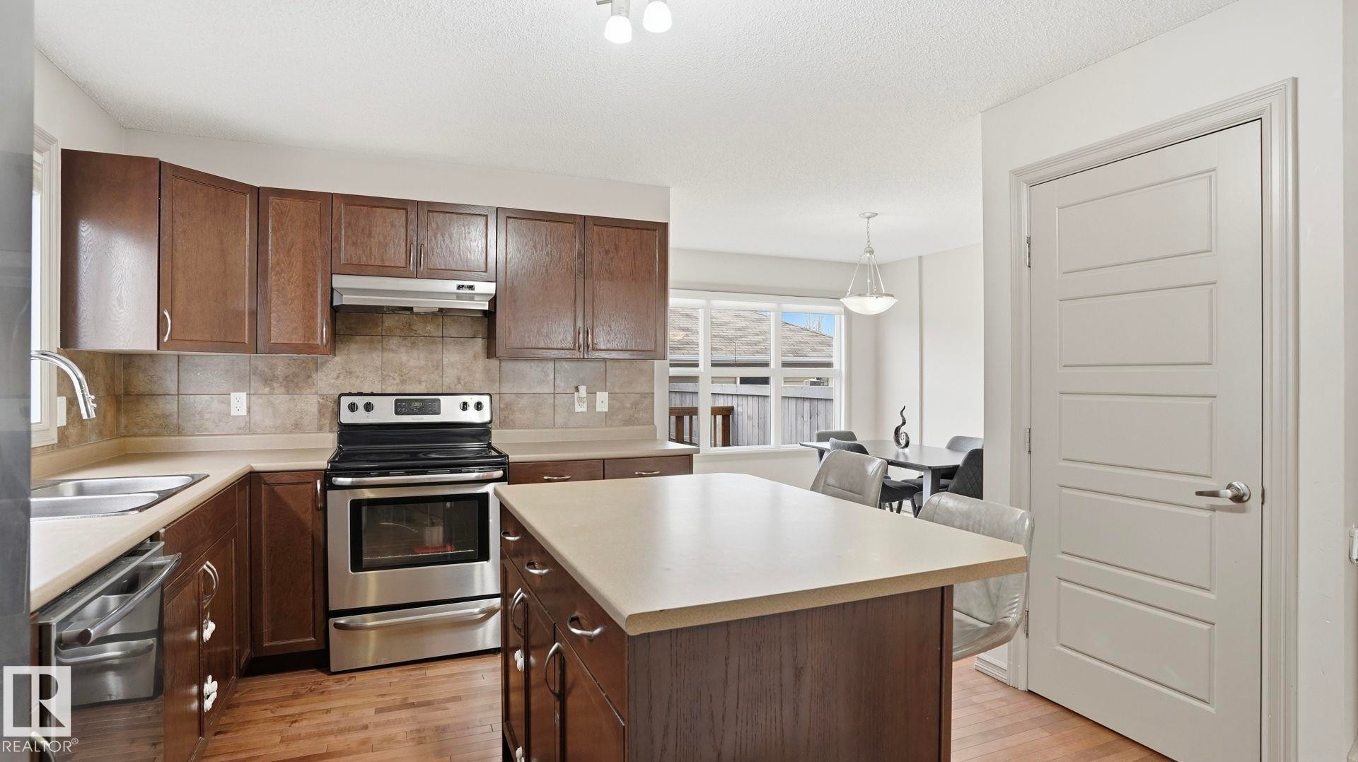 Kitchen featuring wood-finish flooring, dark wood cabinetry, stainless steel appliances, and a central island with a light-toned countertop - 8210 180 Avenue Nw, Edmonton, AB - Indoor Photo Showing Kitchen With Double Sink