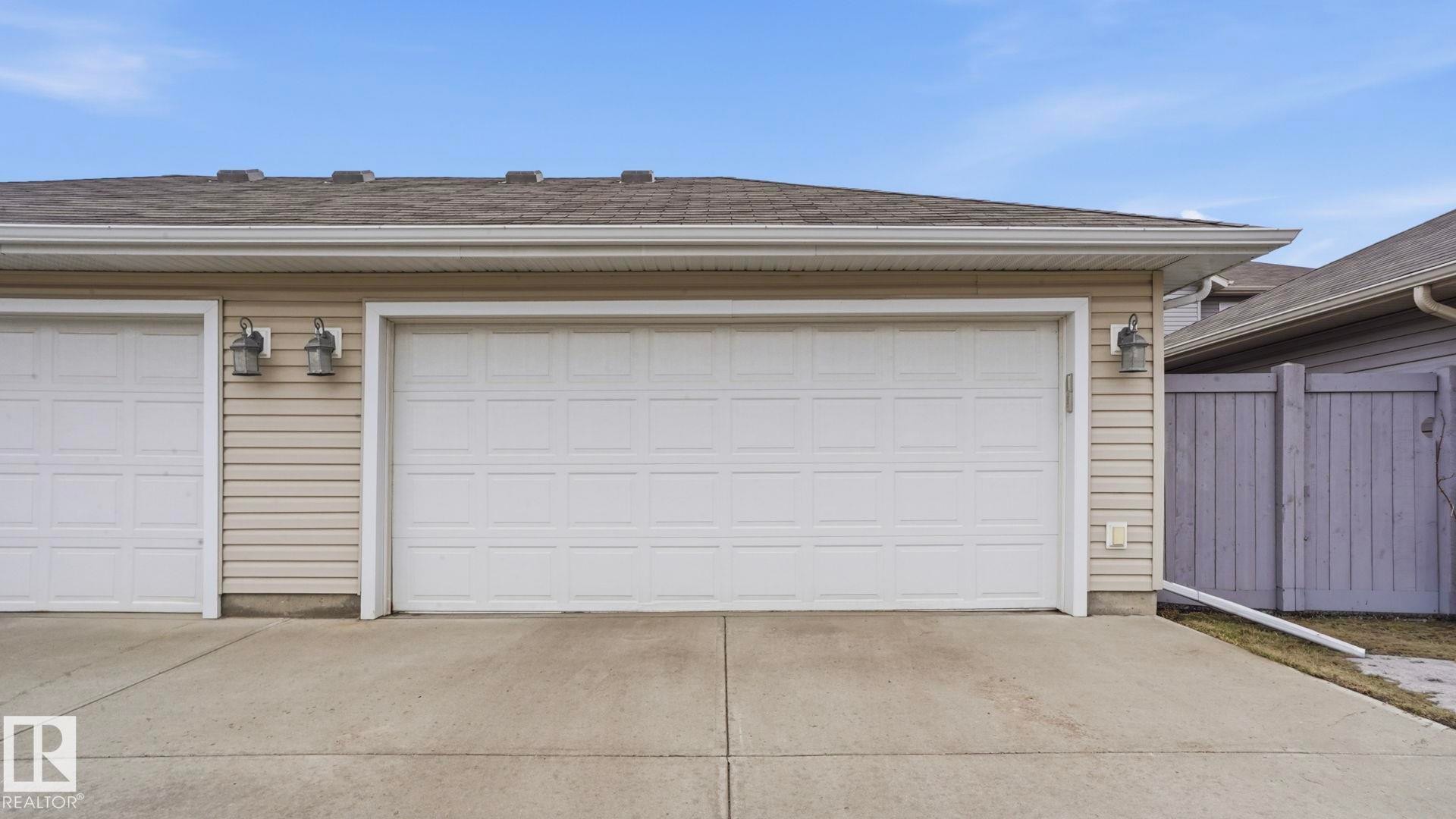 Two white panel garage doors with exterior lighting, beige horizontal siding, and a concrete driveway - 8210 180 Avenue Nw, Edmonton, AB - Outdoor With Exterior