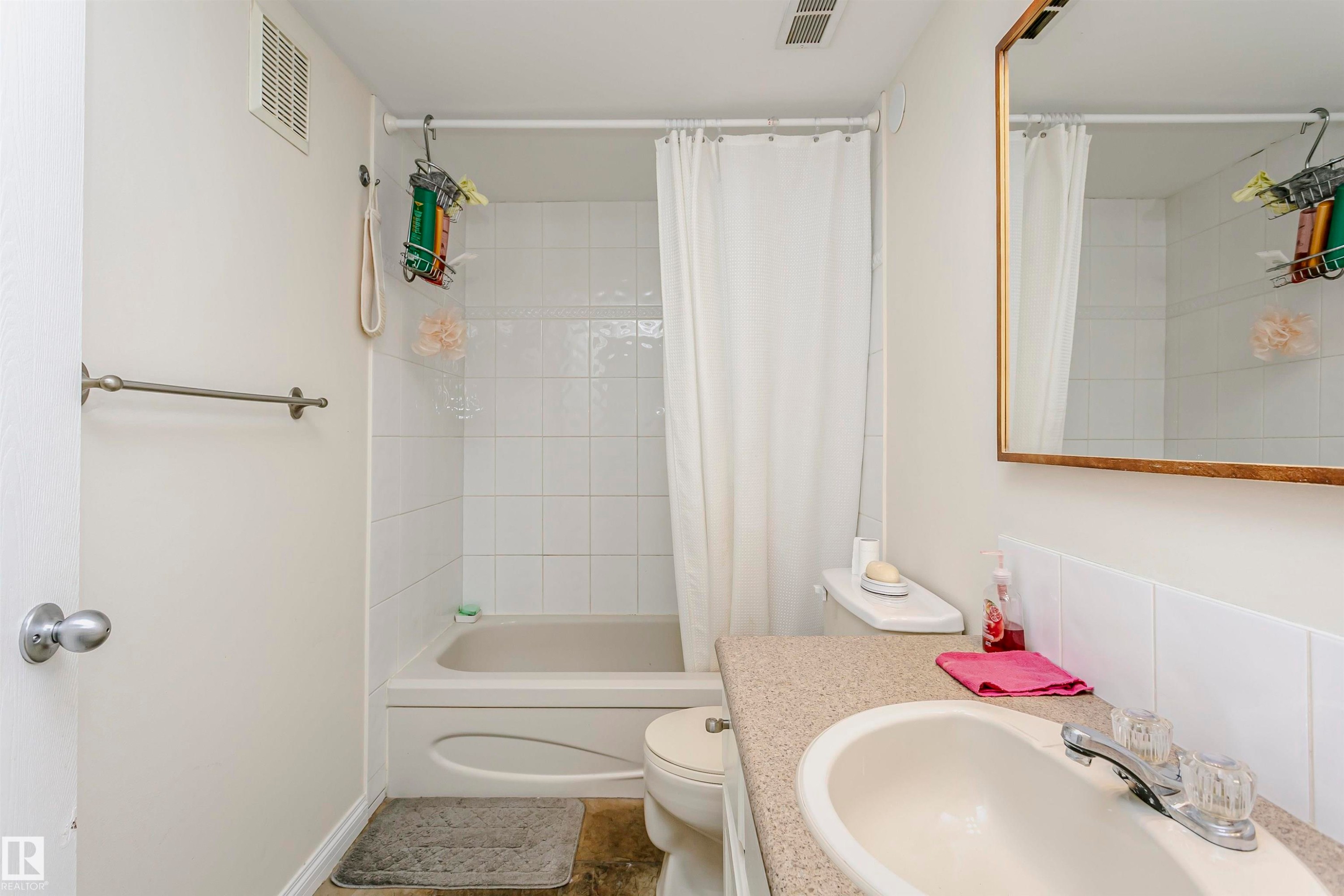 Bathroom featuring a bathtub with white tile surround, a built-in vanity with a composite countertop and integrated sink, and a wall-mounted mirror with a gold-tone frame - #303 10026 112 Street Nw, Edmonton, AB - Indoor Photo Showing Bathroom
