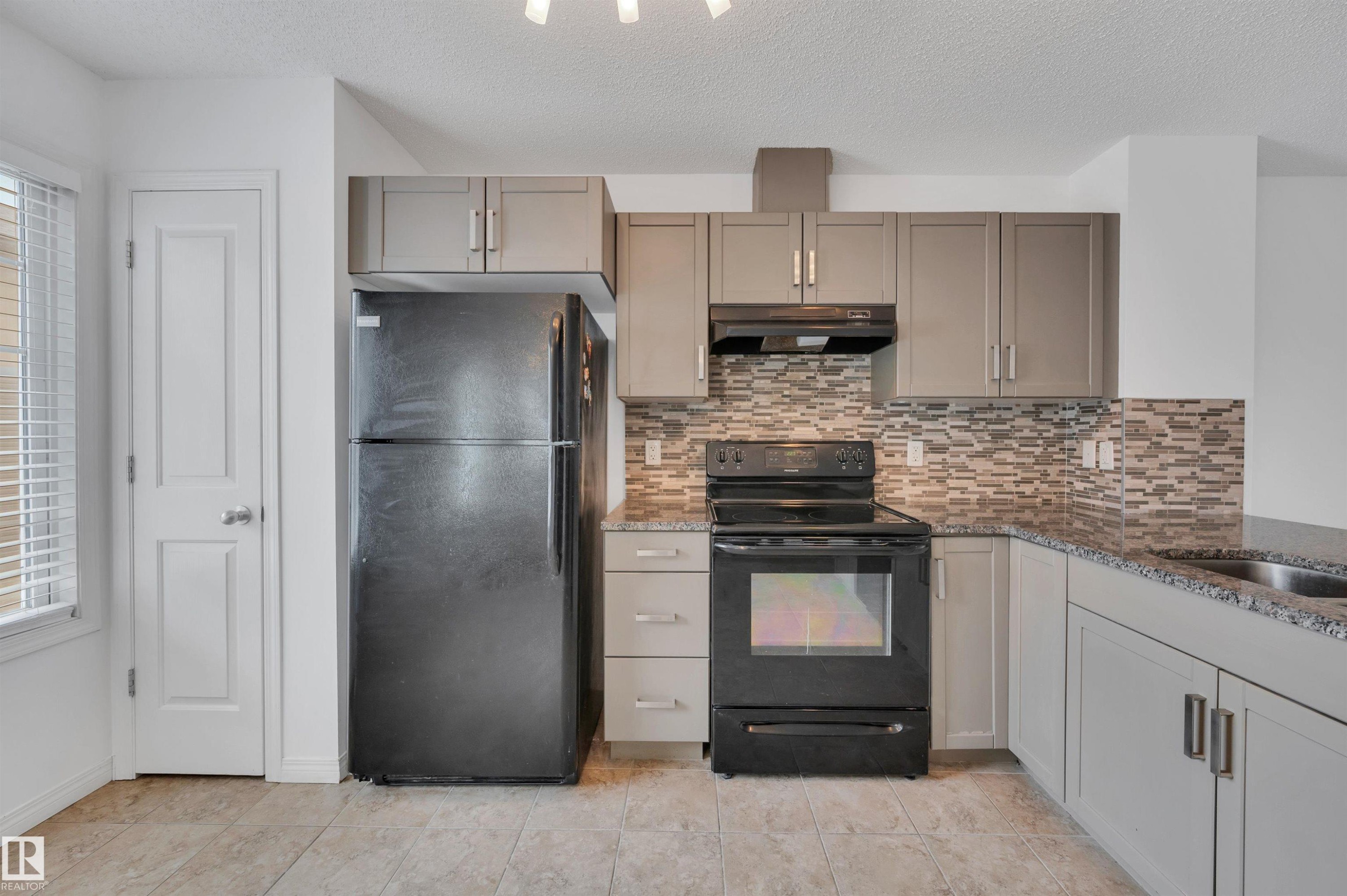 Kitchen featuring light gray cabinetry, dark granite-look countertops, a mosaic tile backsplash, black appliances, and neutral tile flooring - 13 1030 Chappelle Boulevard, Edmonton, AB - Indoor Photo Showing Kitchen