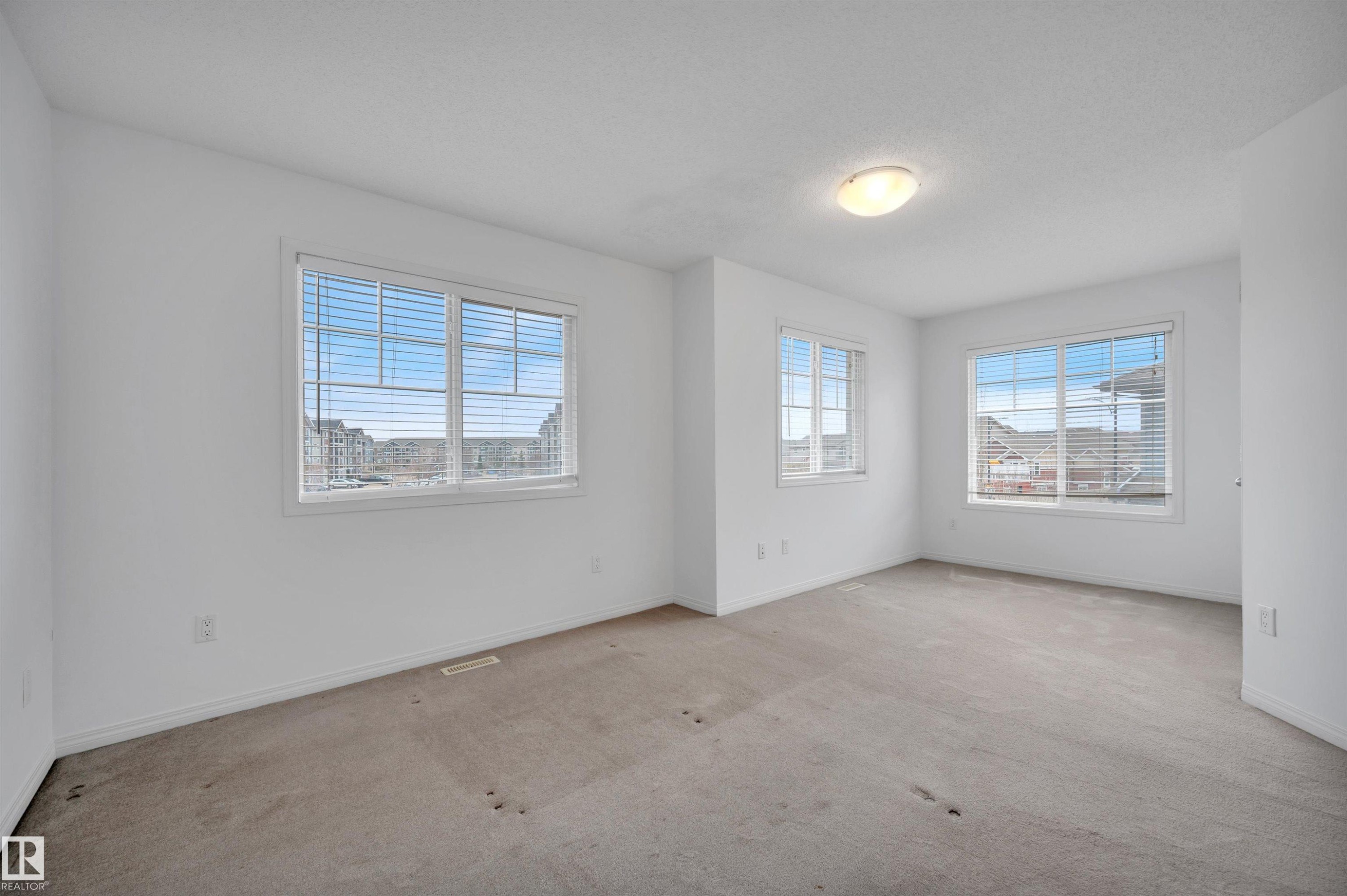 Spacious room featuring multiple windows with blinds, light-colored carpeting, white baseboards, and a flush-mount ceiling light fixture - 13 1030 Chappelle Boulevard, Edmonton, AB - Indoor Photo Showing Other Room