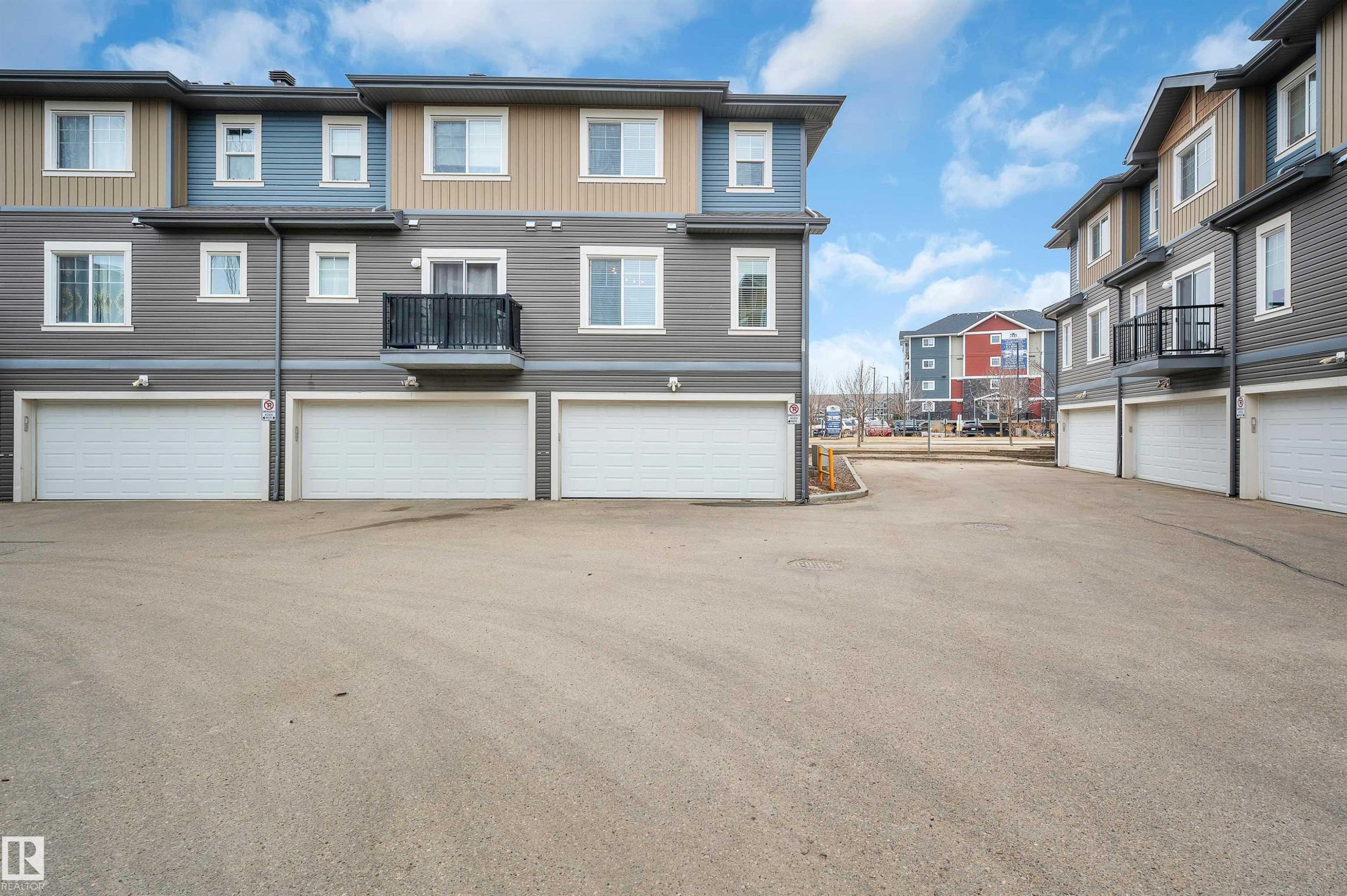 Multi-level townhome exterior featuring horizontal siding in shades of grey, blue, and tan, individual white garage doors, and a private balcony with black metal railings - 13 1030 Chappelle Boulevard, Edmonton, AB - Outdoor With Facade