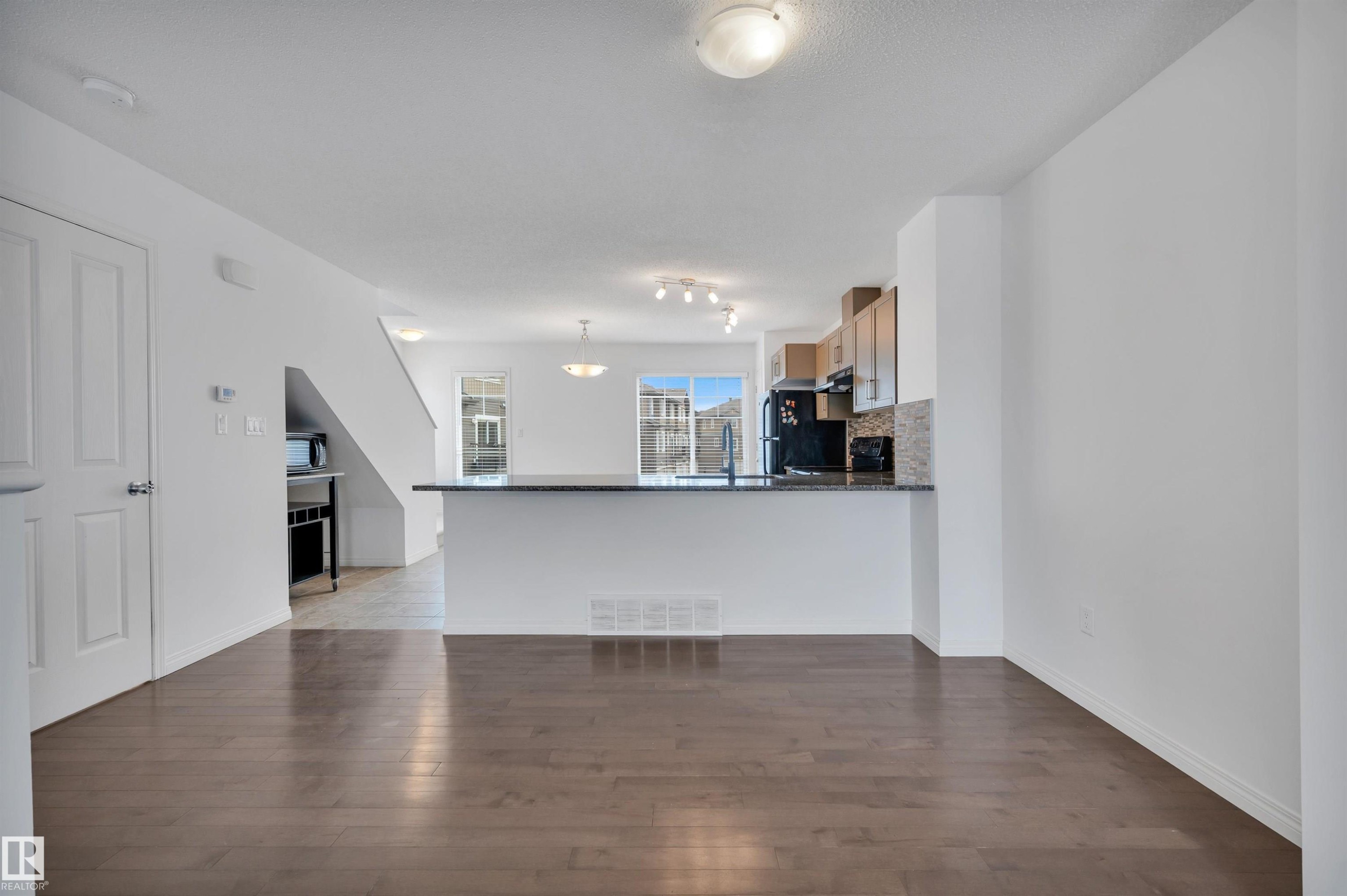 Spacious open-concept living area featuring wood-finish flooring, a kitchen island with a dark countertop, light wood cabinetry, and a mosaic tile backsplash - 13 1030 Chappelle Boulevard, Edmonton, AB - Indoor