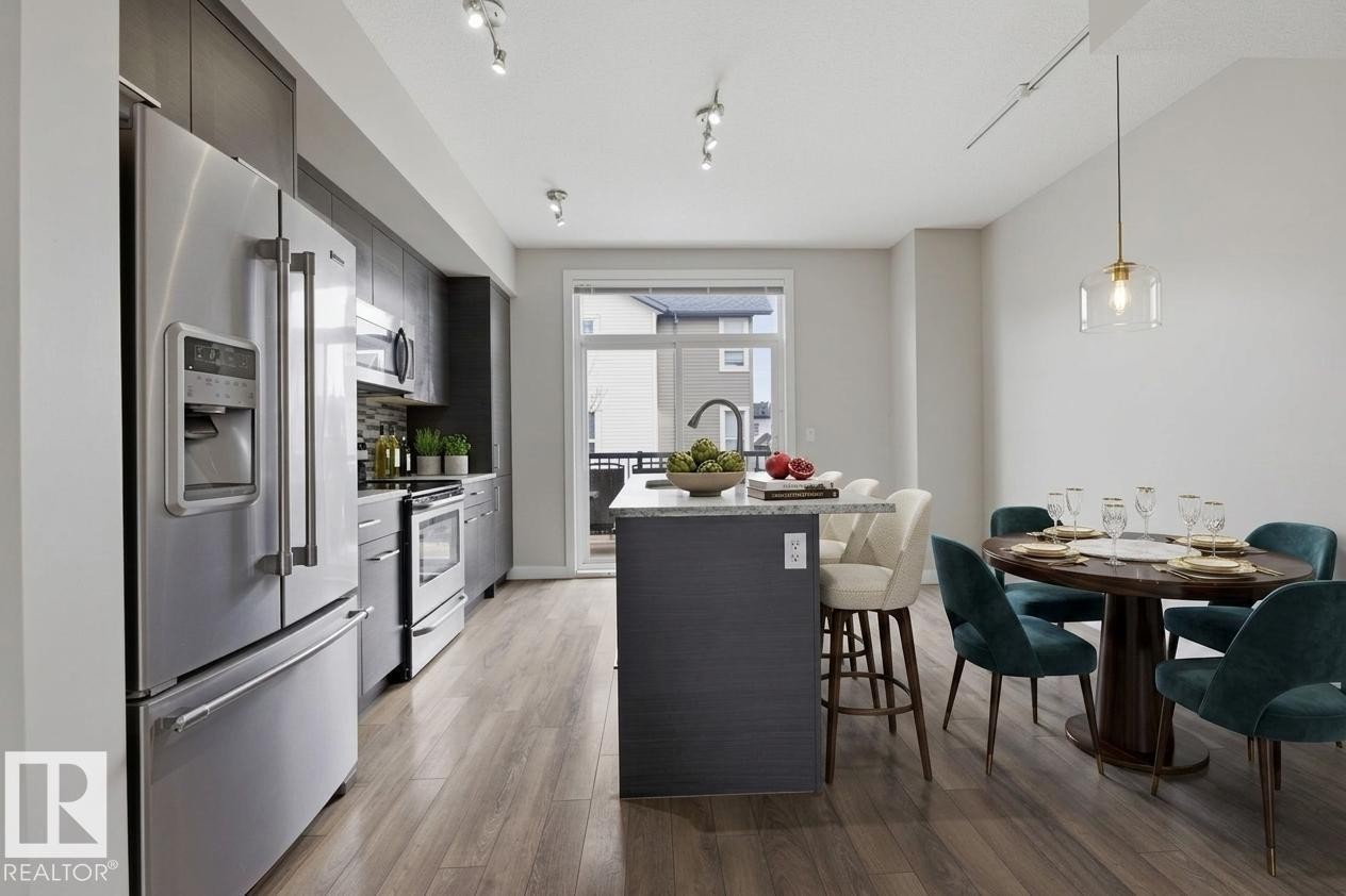Contemporary kitchen and dining area featuring stainless steel appliances, dark cabinetry, and wood-finish flooring - 18, 2560 Pegasus Boulevard, Edmonton, AB - Indoor