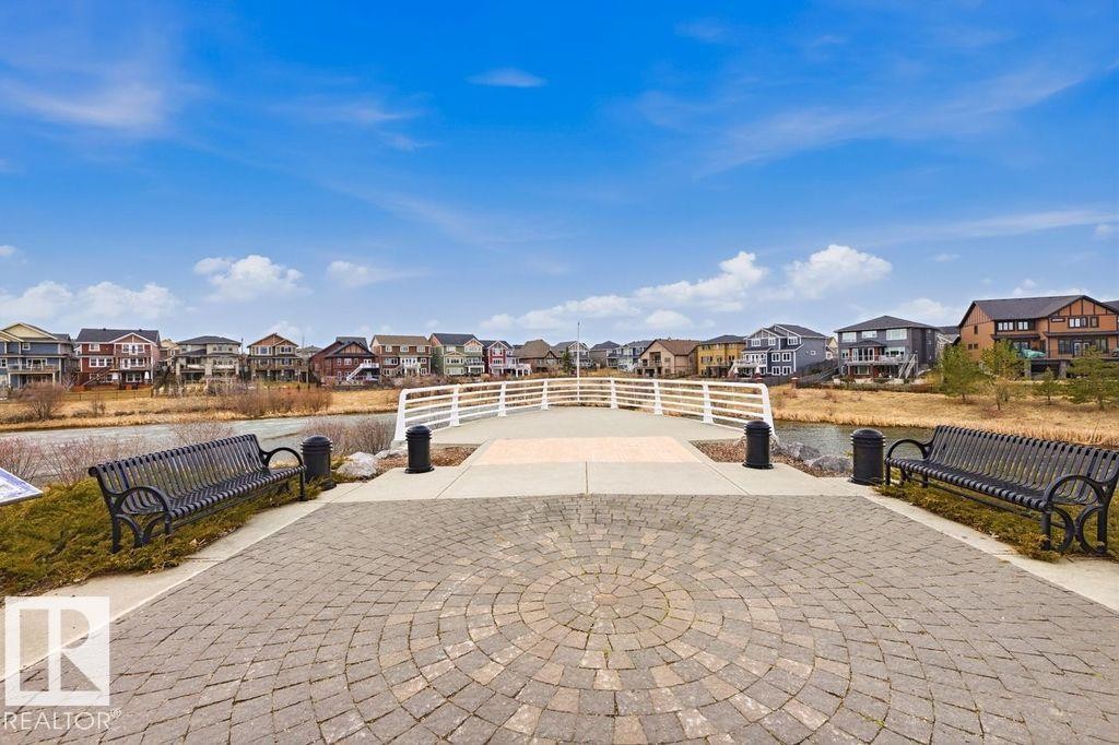Paved walkway featuring a circular paver design, two black metal benches, and a white pedestrian bridge over a body of water - 18, 2560 Pegasus Boulevard, Edmonton, AB - Outdoor With View
