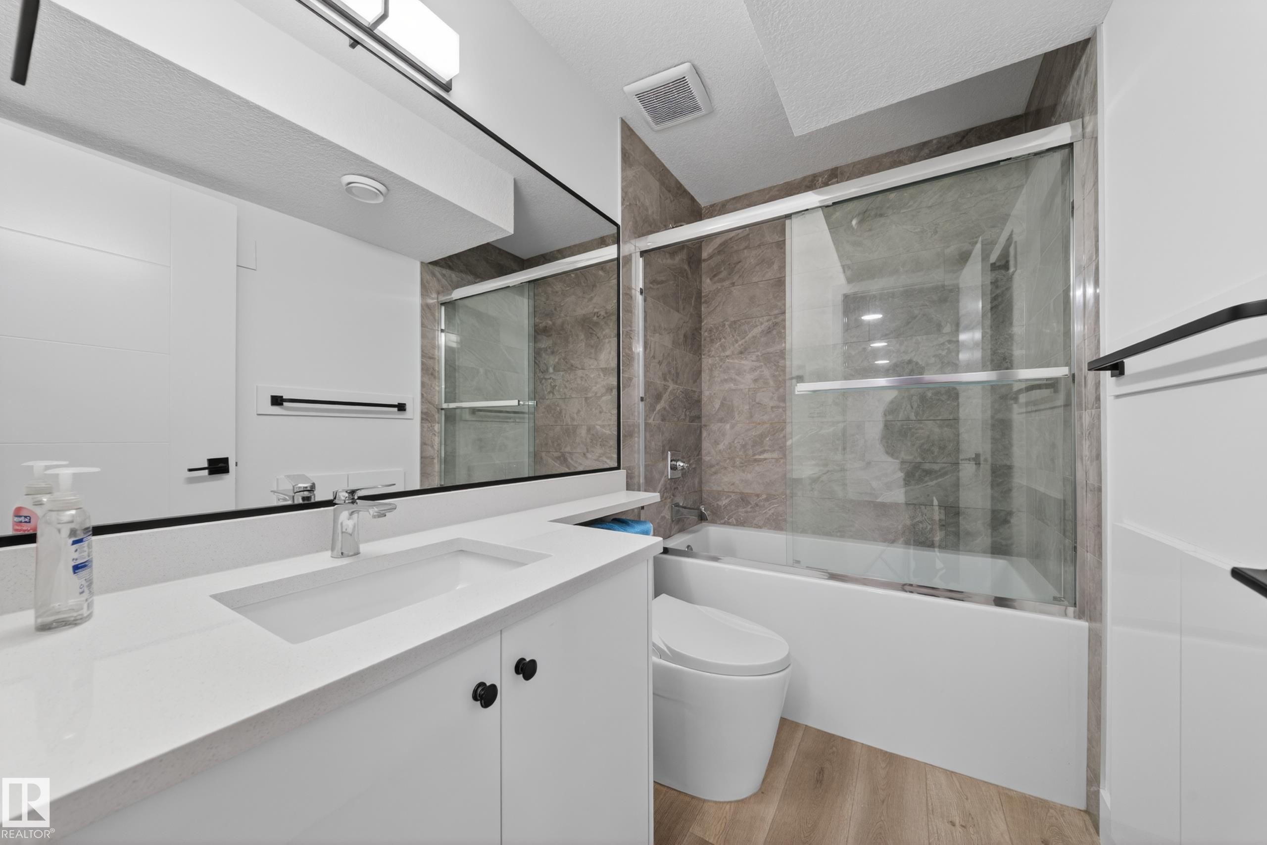 Modern bathroom featuring a white vanity with integrated sink, a large wall-mounted mirror, a bathtub with glass sliding doors, and wood-finish flooring - 7414 Chivers Crescent Sw, Edmonton, AB - Indoor Photo Showing Bathroom