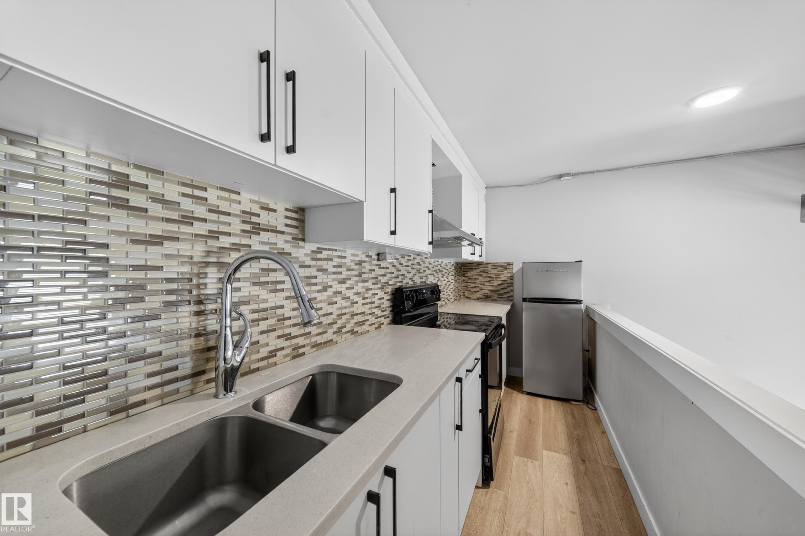 Contemporary kitchen featuring white cabinetry with black hardware, a stainless steel double basin sink, a pull-down faucet, and light wood-finish flooring - 7414 Chivers Crescent Sw, Edmonton, AB - Indoor Photo Showing Kitchen With Double Sink