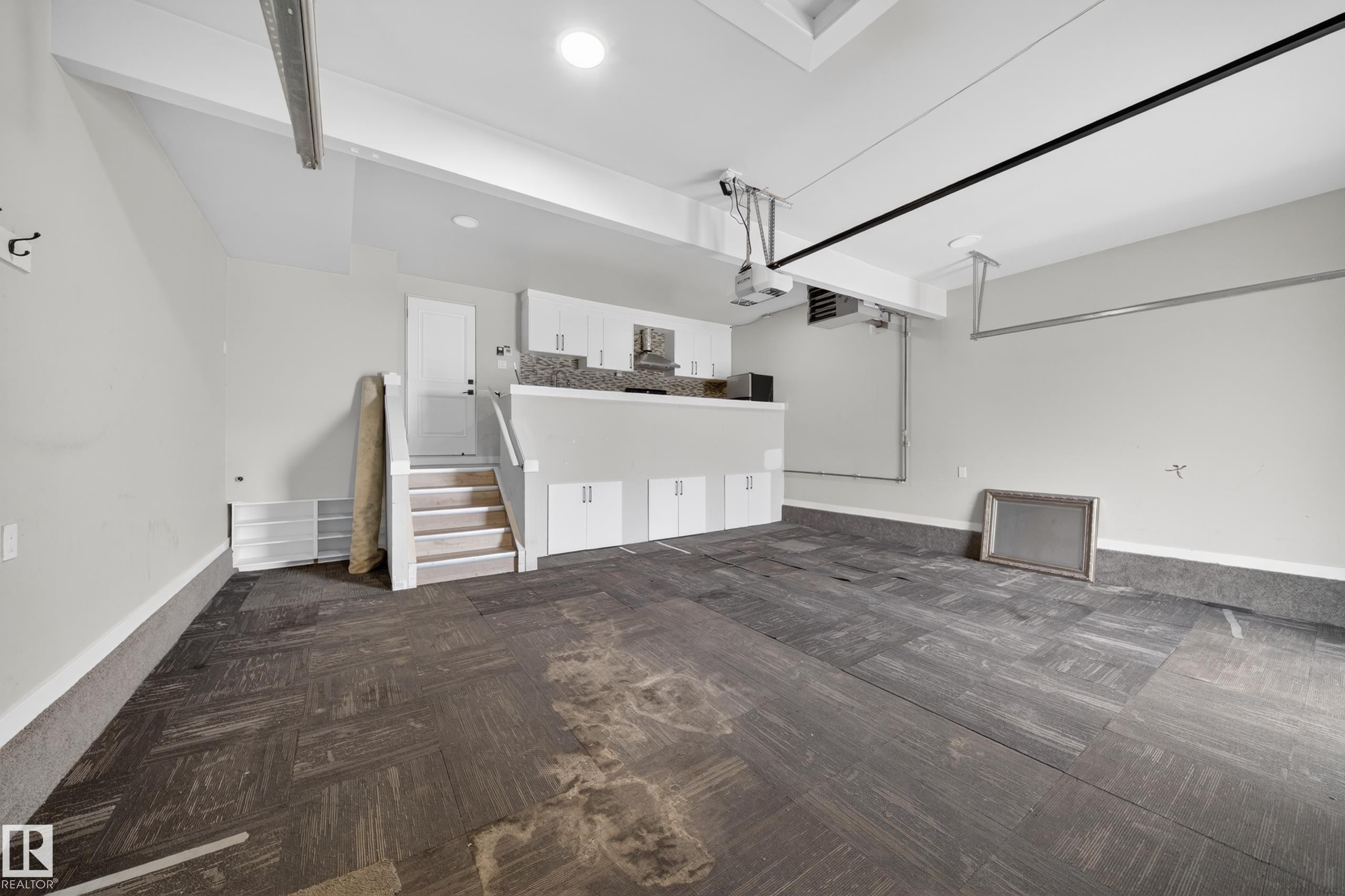 Spacious garage featuring dark wood-finish flooring, overhead lighting, and an integrated kitchenette with white cabinetry, a stone-look backsplash, and a built-in microwave - 7414 Chivers Crescent Sw, Edmonton, AB - Indoor Photo Showing Other Room