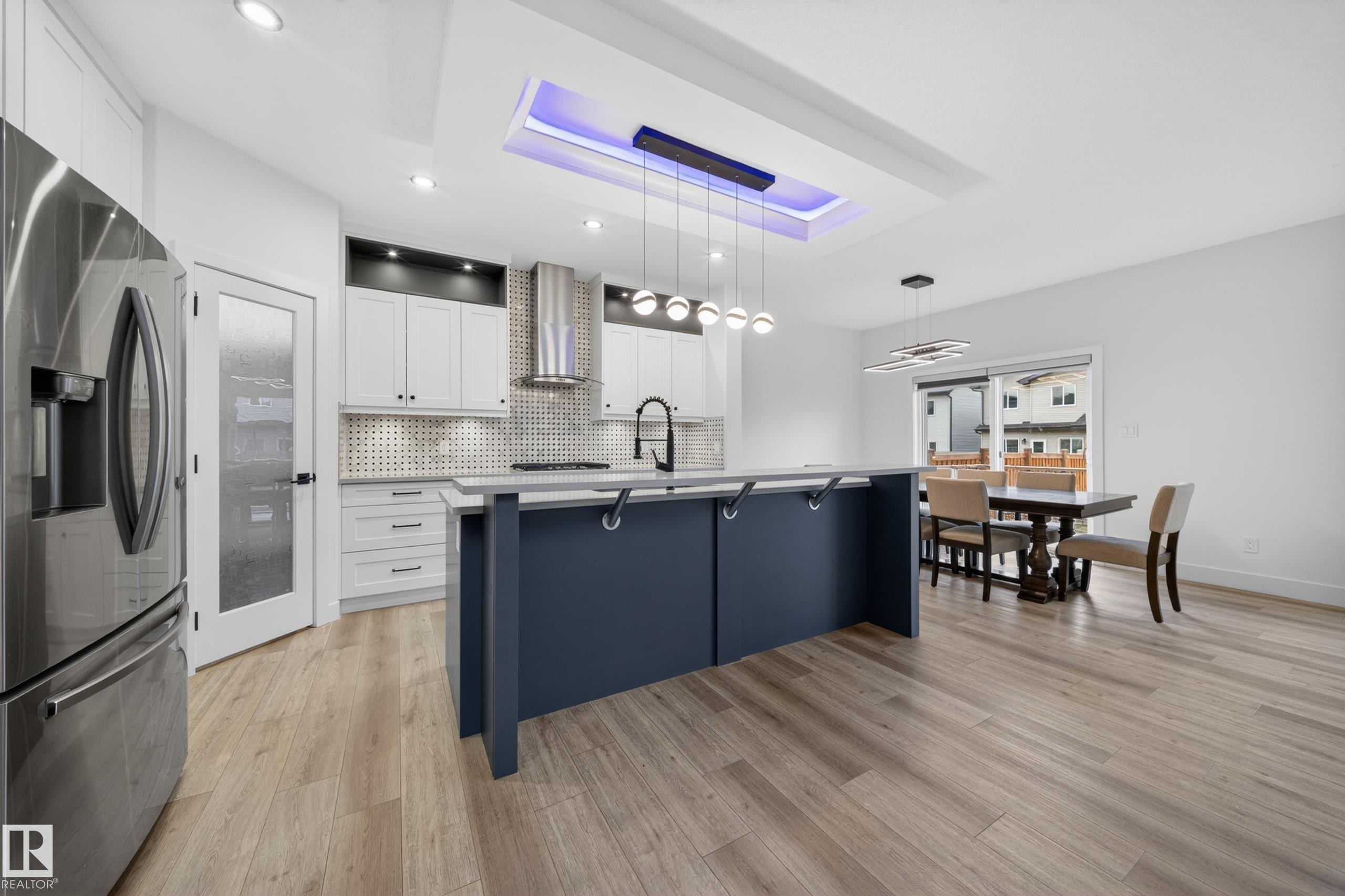 Open-concept kitchen and dining area featuring light wood-finish flooring, a kitchen island with a built-in sink, white cabinetry with a patterned backsplash, stainless steel appliances, and a tray ceiling with recessed lighting - 7414 Chivers Crescent Sw, Edmonton, AB - Indoor Photo Showing Kitchen With Upgraded Kitchen