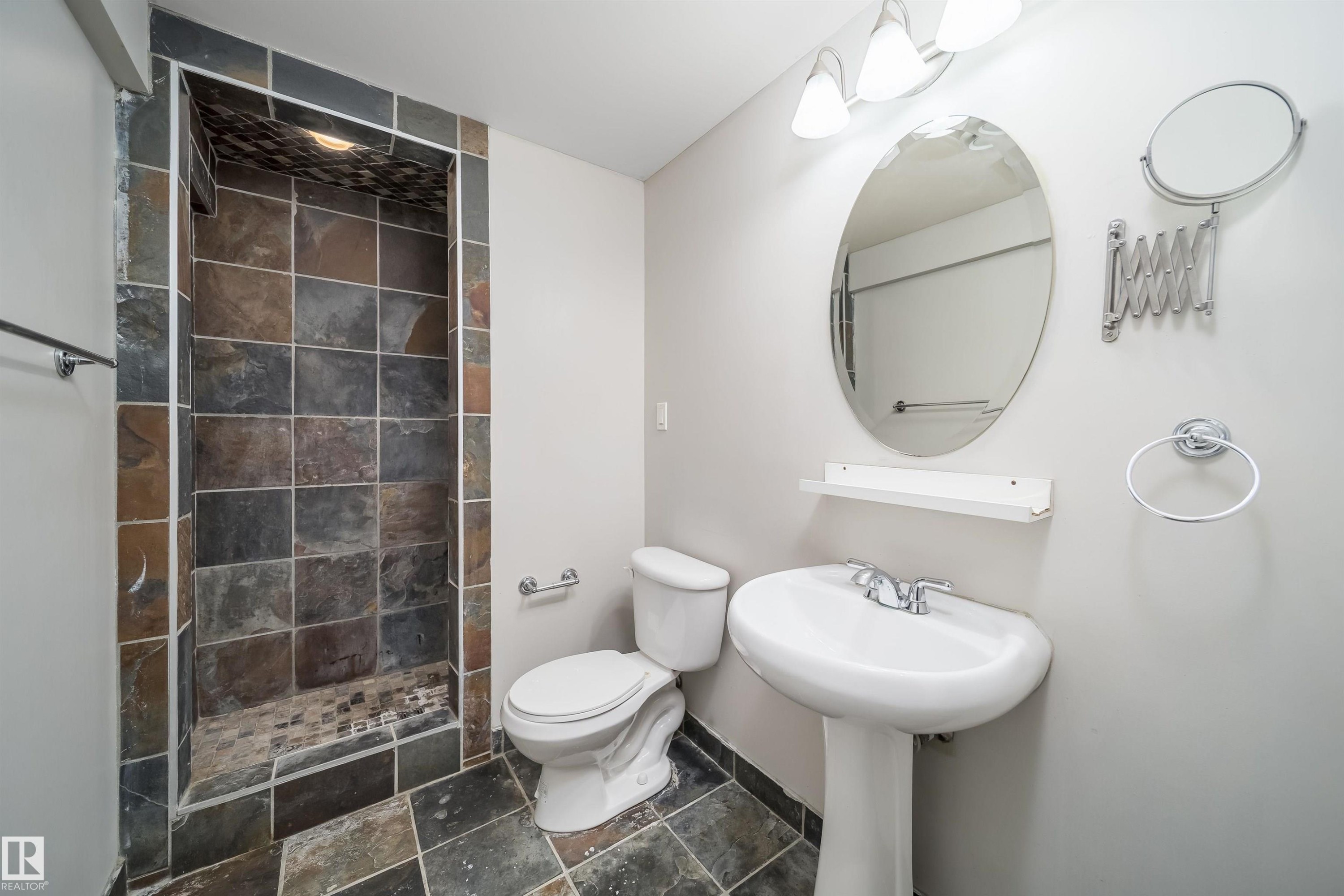 Walk-in shower featuring multi-toned slate tile, a coordinating slate tile floor, a pedestal sink with chrome faucet, an oval vanity mirror, and three-light vanity fixture - 50 14603 Miller Boulevard, Edmonton, AB
