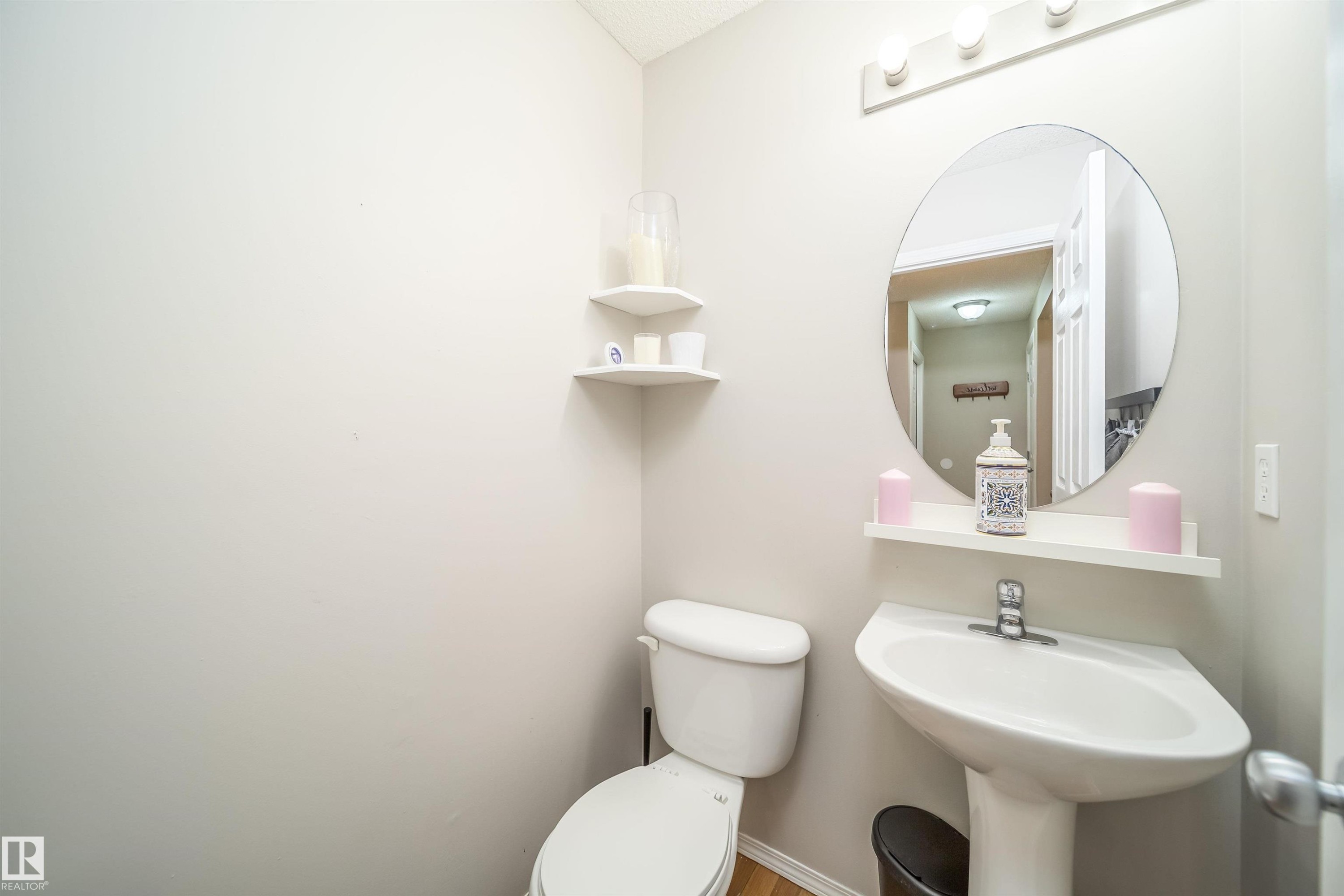 Powder room featuring a pedestal sink, round wall mirror, and white wall-mounted shelving - 50 14603 Miller Boulevard, Edmonton, AB