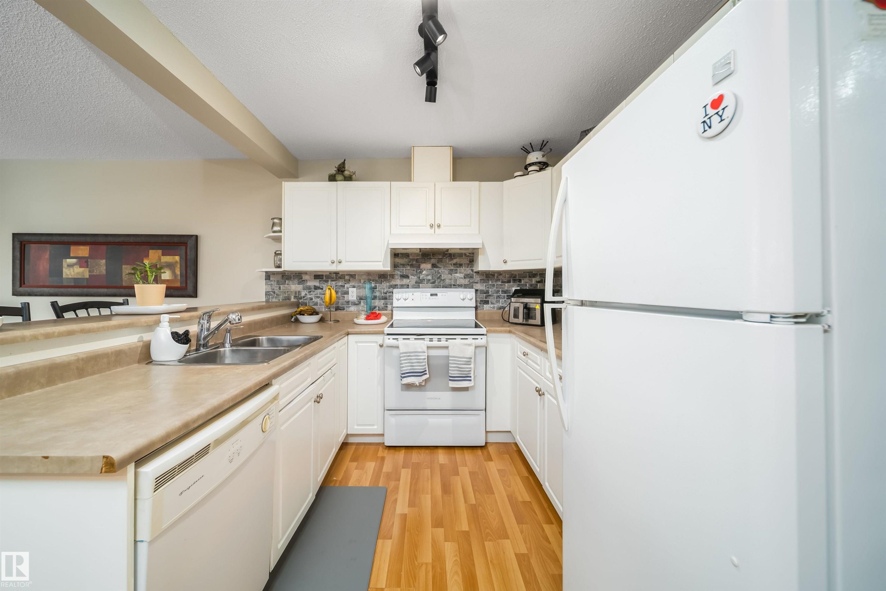 White kitchen featuring wood-finish flooring, light-toned countertops, and a mosaic tile backsplash - 50 14603 Miller Boulevard, Edmonton, AB