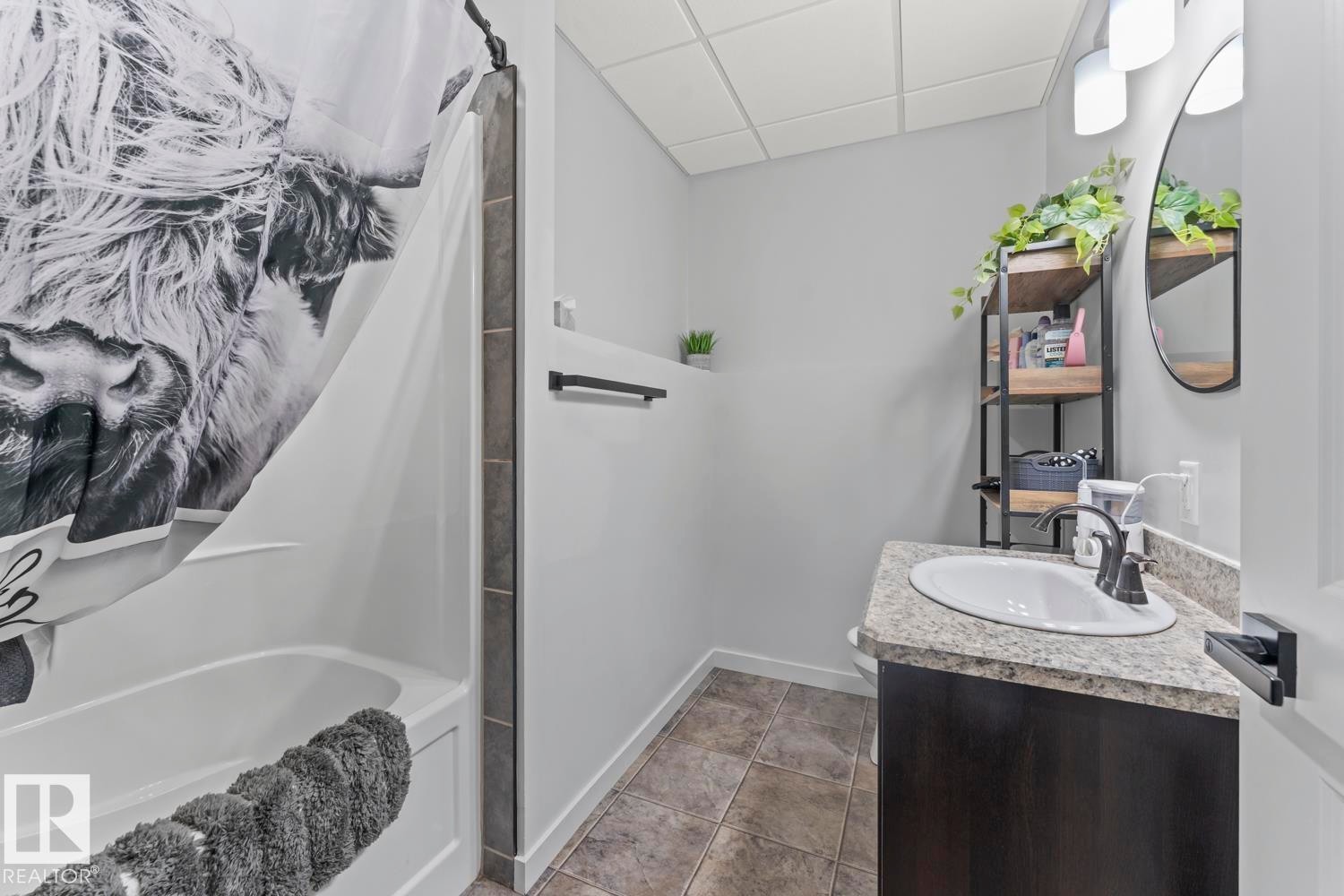 Bathroom featuring a white bathtub, tiled shower surround, dark wood-finish vanity with a light speckled countertop, and a circular wall mirror with a black frame - 4926 58 Ave, Cold Lake, AB - Indoor Photo Showing Bathroom
