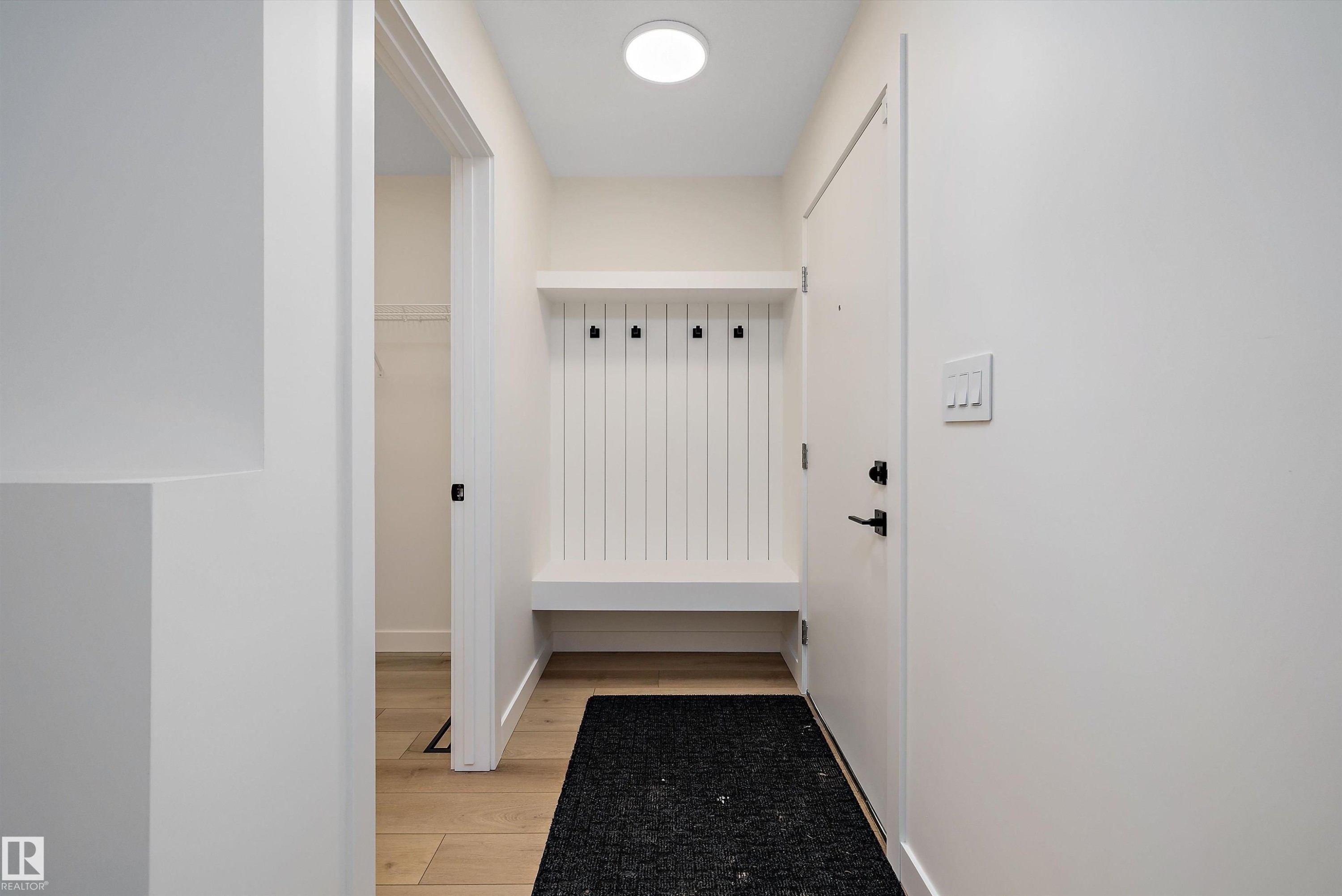 Entryway mudroom bench with integrated coat hooks, showcasing wood-finish flooring, a contemporary flush-mount ceiling light, and a white interior door with matte black hardware - 11418 96 Street, Edmonton, AB - Indoor Photo Showing Other Room