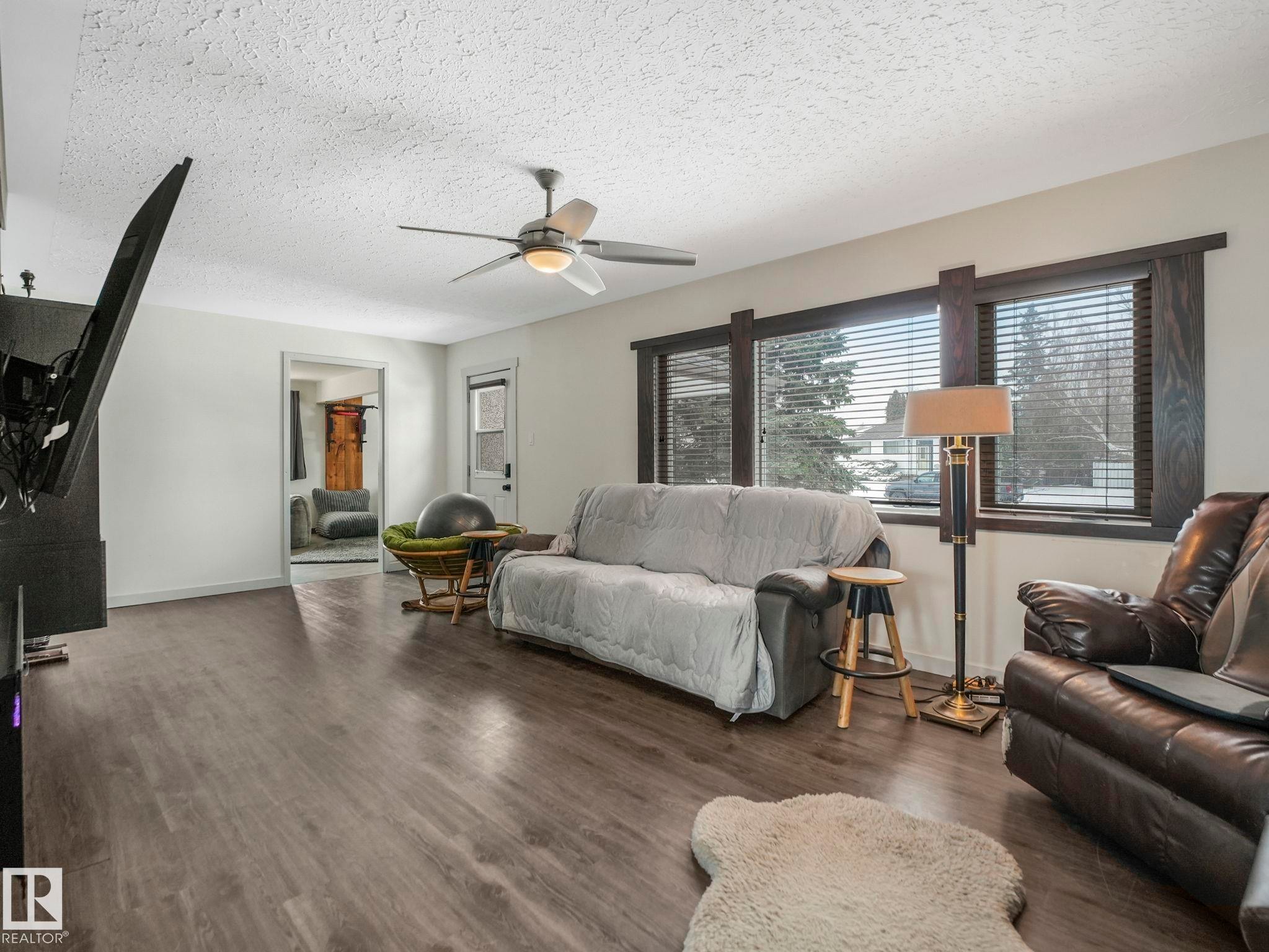 Spacious living area featuring wood-finish flooring, a ceiling fan with integrated lighting, and multiple windows with dark wood trim - 7607 70 Avenue, Edmonton, AB - Indoor Photo Showing Living Room