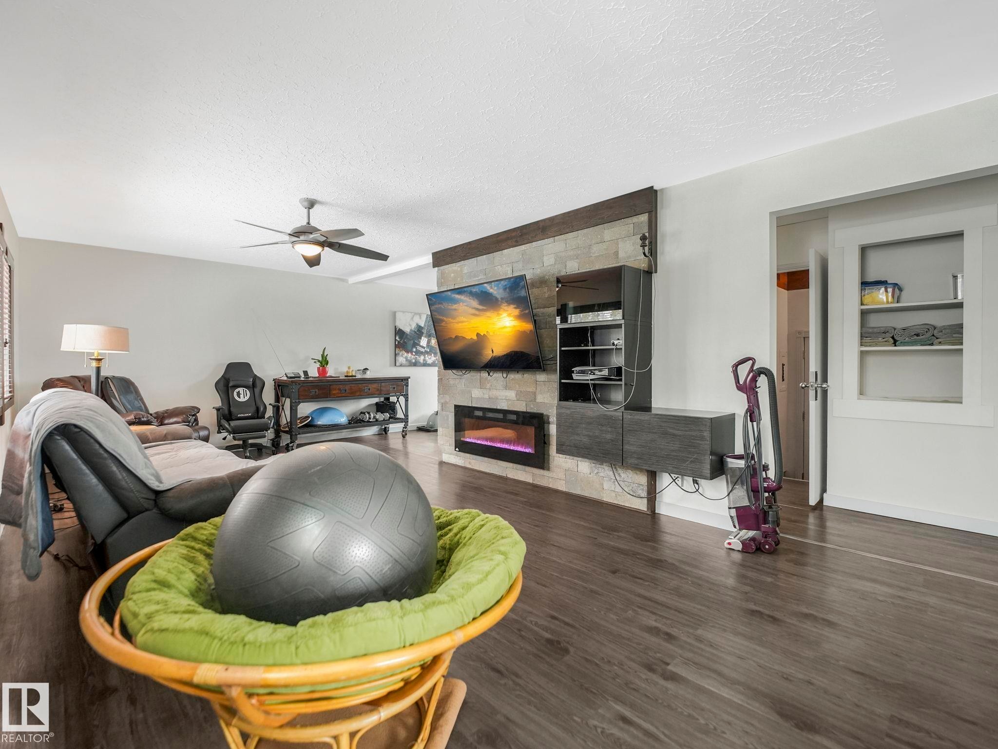 Living area featuring wood-finish flooring, a ceiling fan with integrated lighting, and a stone-look accent wall with an electric fireplace - 7607 70 Avenue, Edmonton, AB - Indoor With Fireplace