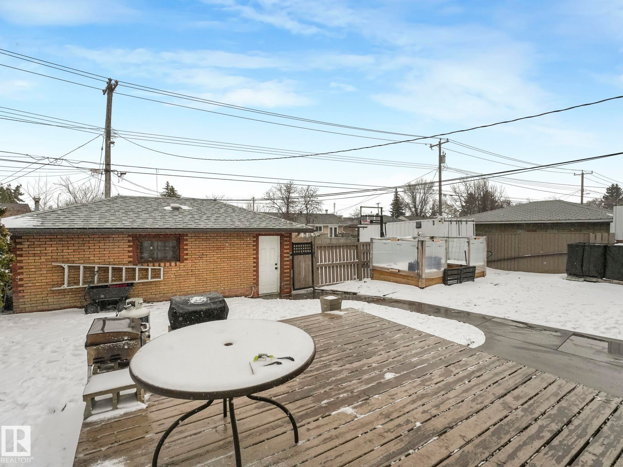 Expansive wood-finish deck with a patio table, accompanied by a brick garage with a shingle roof and a separate white door - 7607 70 Avenue, Edmonton, AB - Outdoor With Deck Patio Veranda