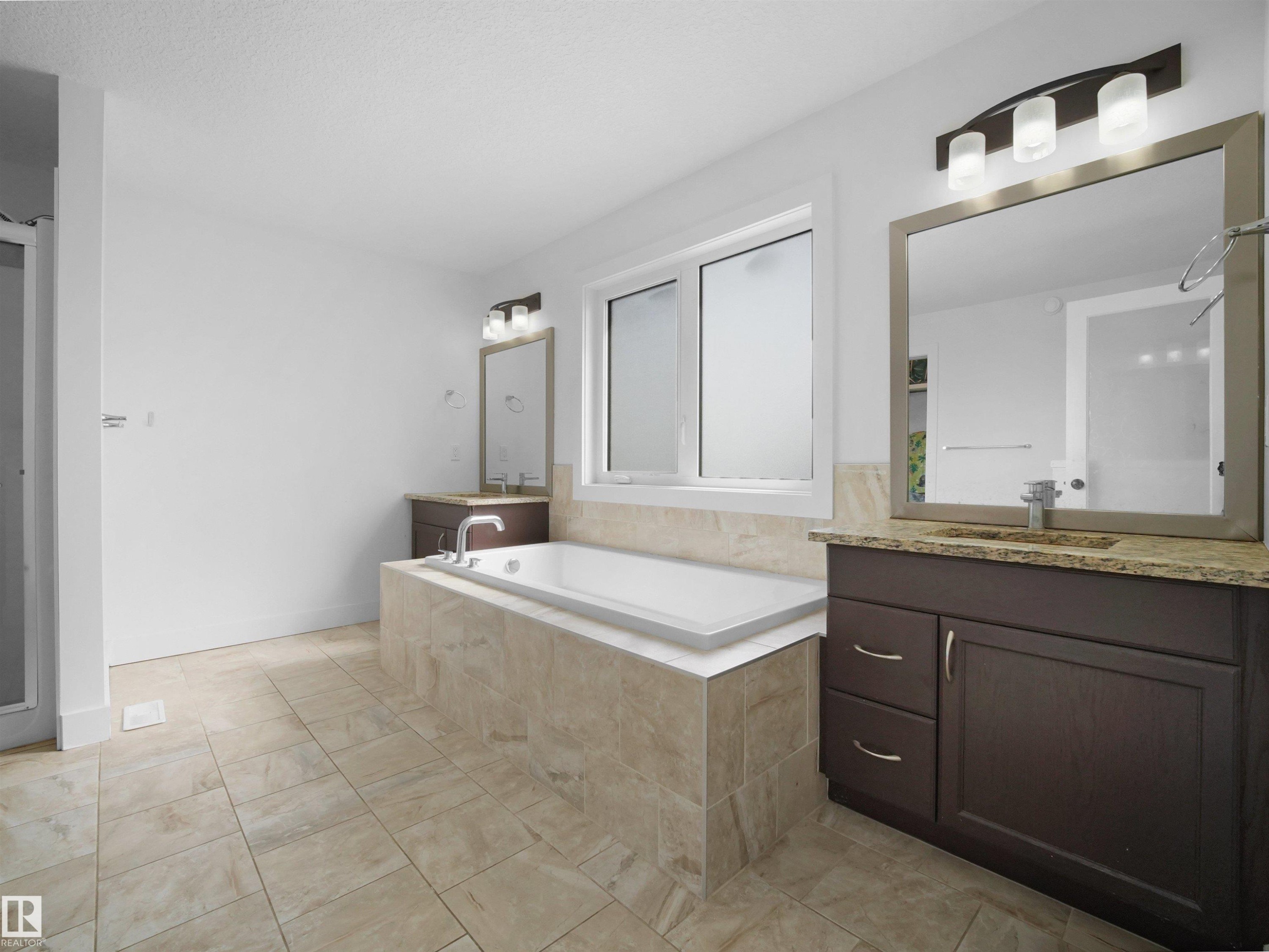 Bathroom featuring a built-in bathtub with tiled surround, a window with frosted glass, and two separate vanity areas with granite countertops and mirrors - 4231 Charles Close, Edmonton, AB - Indoor Photo Showing Bathroom