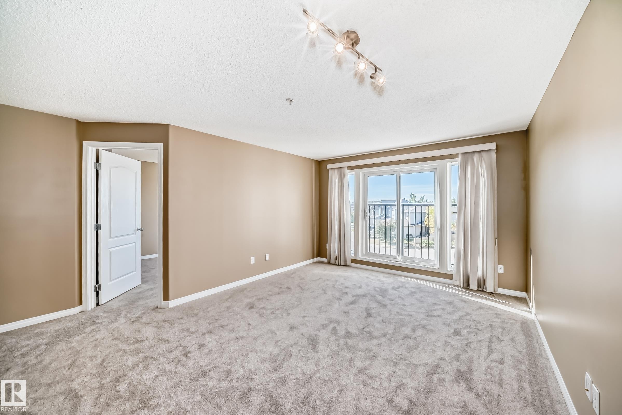 Spacious room featuring neutral paint tones, plush gray carpet flooring, a large sliding glass door with a balcony, track lighting, and a white interior door - 344 1520 Hammond Gate Gate, Edmonton, AB - Indoor Photo Showing Other Room