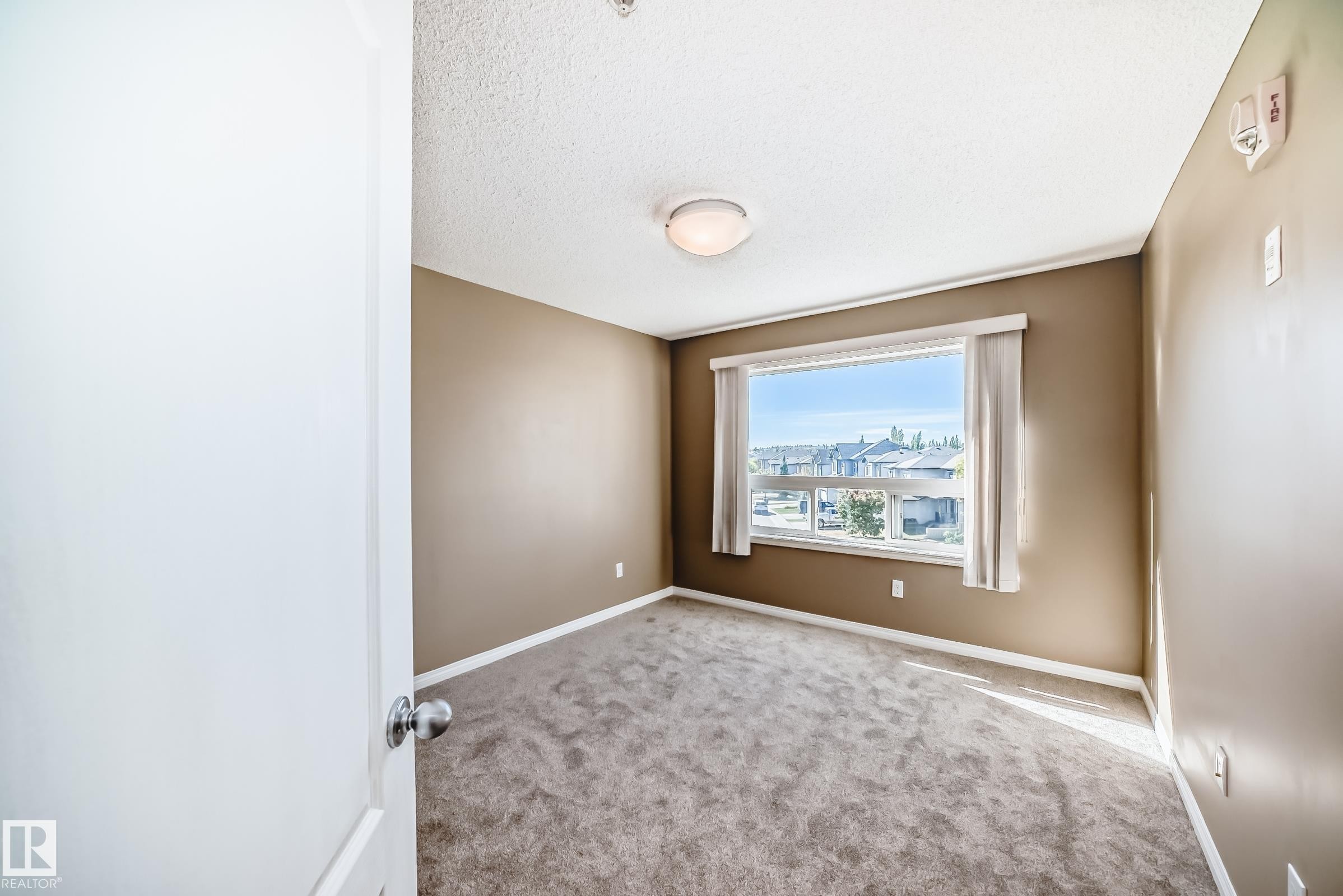 Carpeted room featuring a large window, neutral wall paint, white trim, and a ceiling-mounted light fixture - 344 1520 Hammond Gate Gate, Edmonton, AB - Indoor Photo Showing Other Room
