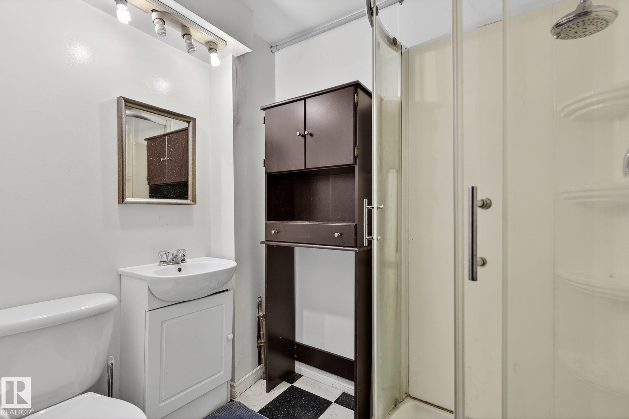 Compact bathroom featuring a glass-enclosed shower, white ceramic toilet, vanity with integrated basin, dark wood-finish storage tower, and checkerboard floor tiling - 9112 130 Avenue, Edmonton, AB - Indoor Photo Showing Bathroom