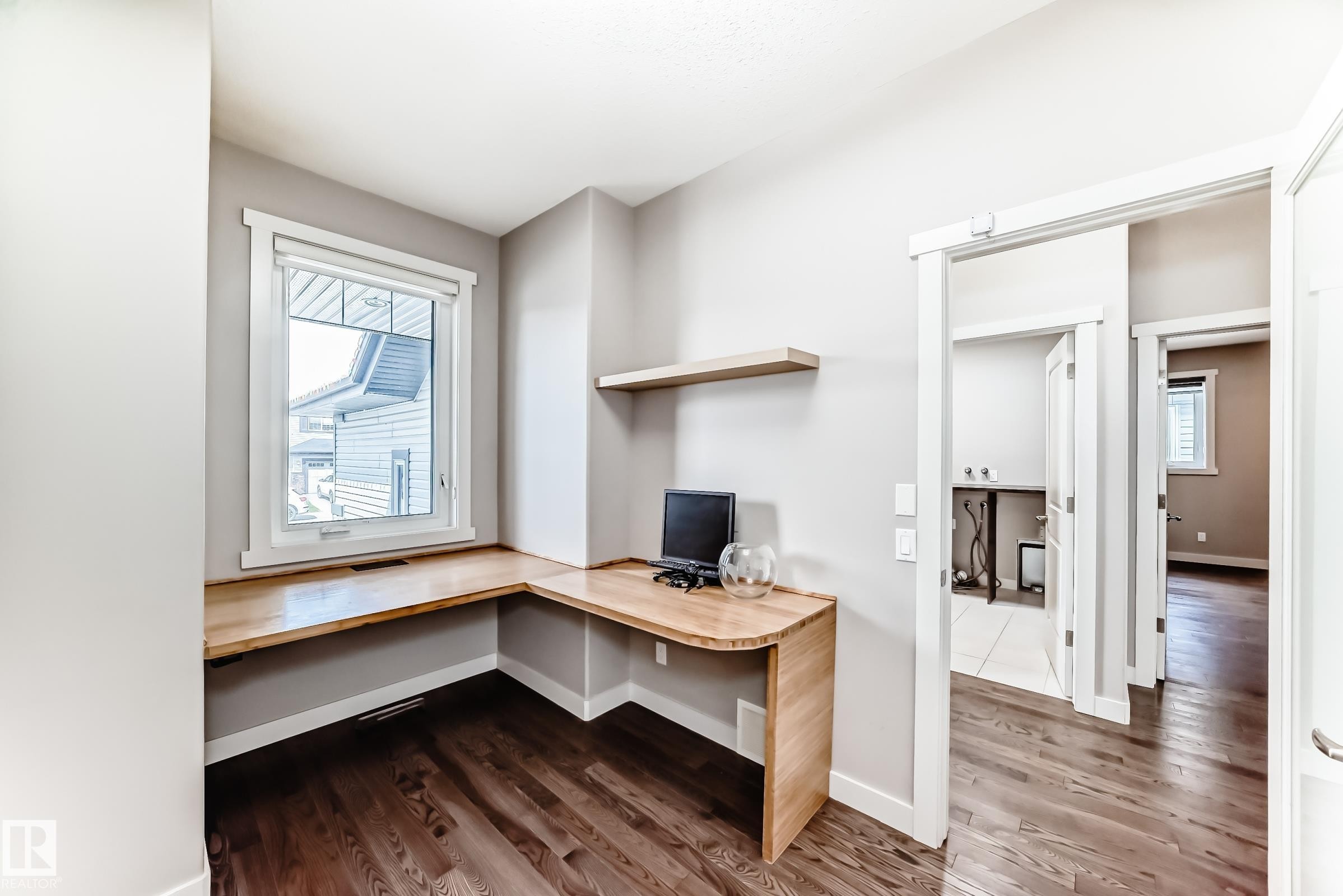 Built-in corner desk with a light wood finish, single floating shelf, single window with white trim, and dark wood-finish flooring - 7719 Getty Wynd, Edmonton, AB