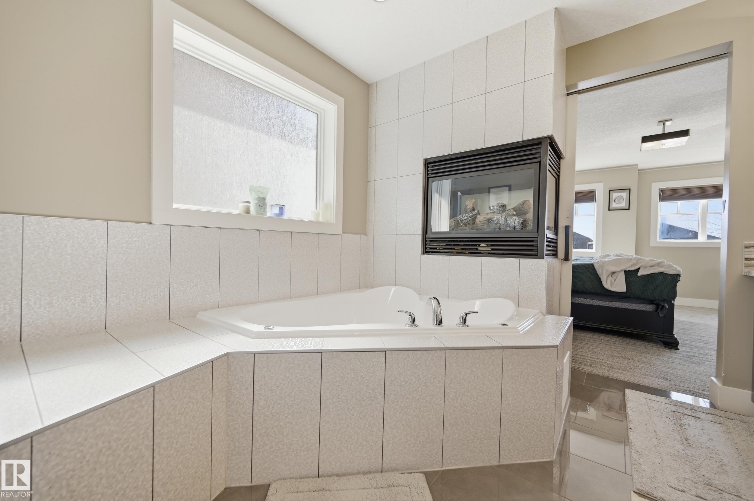 Bathroom featuring a built-in bathtub with chrome fixtures, a window above the tub, and a wall-mounted fireplace - 2018 Armitage Green, Edmonton, AB - Indoor Photo Showing Bathroom
