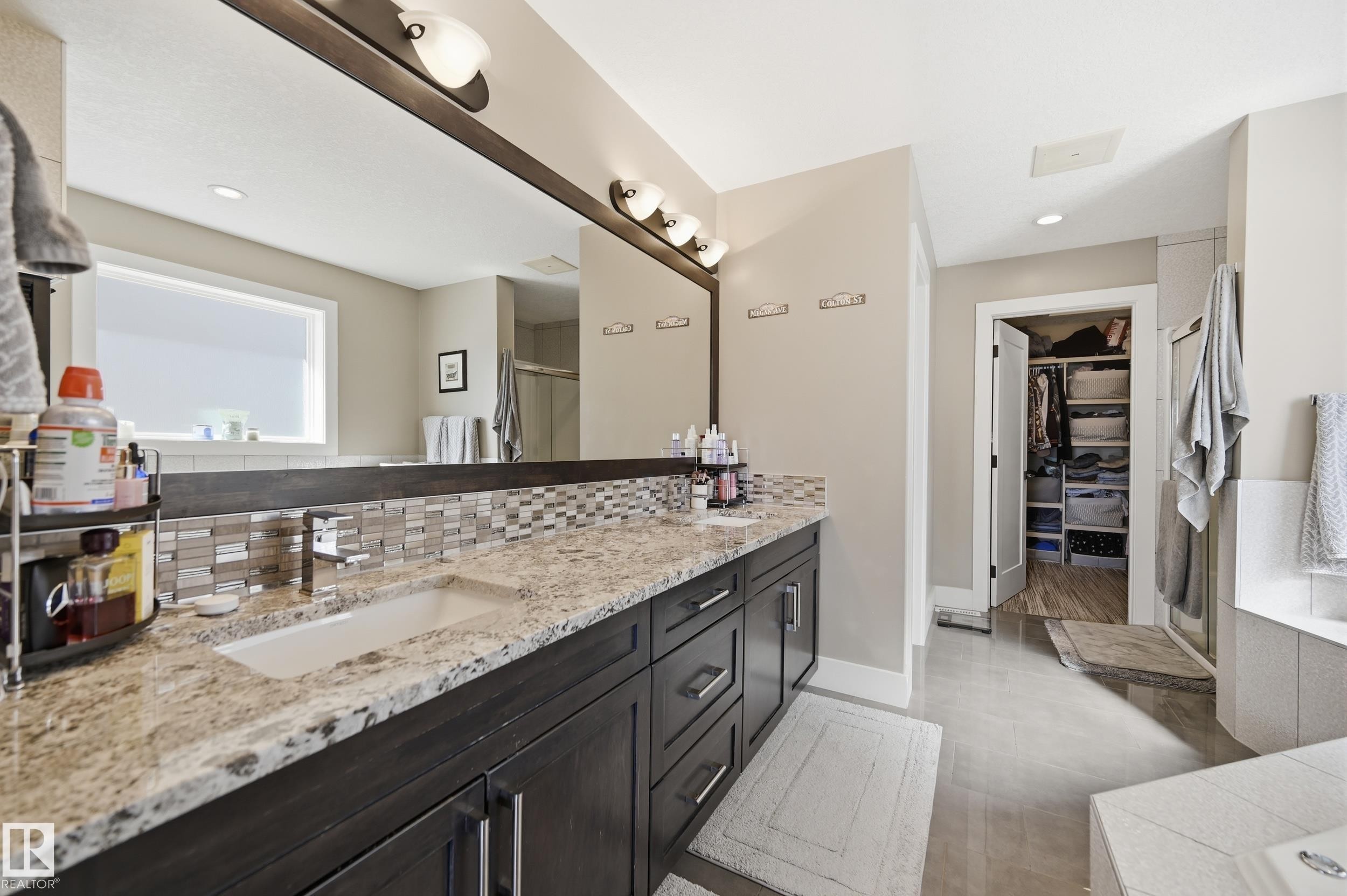 The bathroom features a double vanity with granite countertops, a large mirror with overhead lighting, and a tiled backsplash - 2018 Armitage Green, Edmonton, AB - Indoor Photo Showing Bathroom