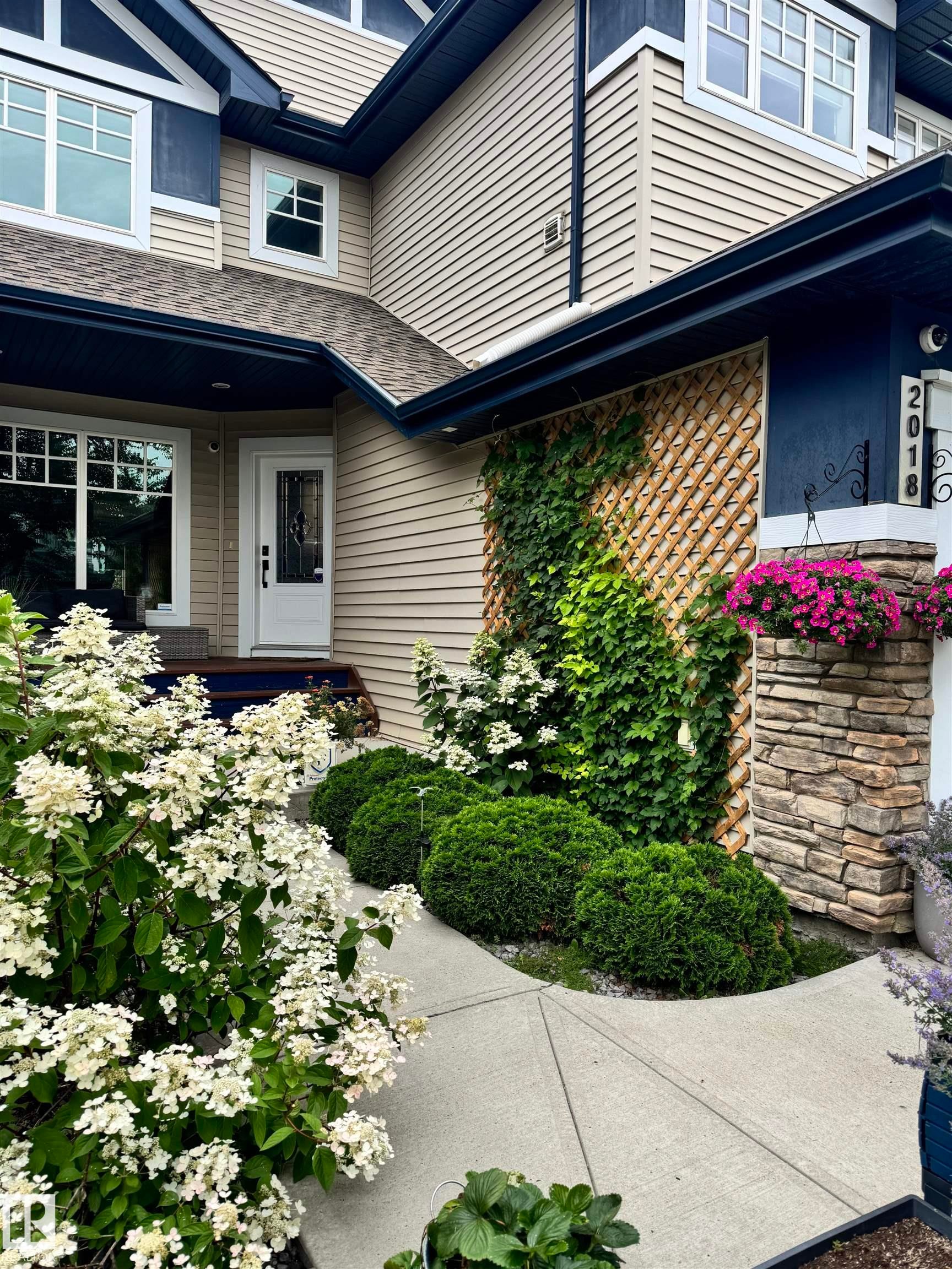 The property features a concrete walkway leading to the entrance, surrounded by well-maintained landscaping including flowering shrubs and neatly trimmed bushes - 2018 Armitage Green, Edmonton, AB - Outdoor With Deck Patio Veranda