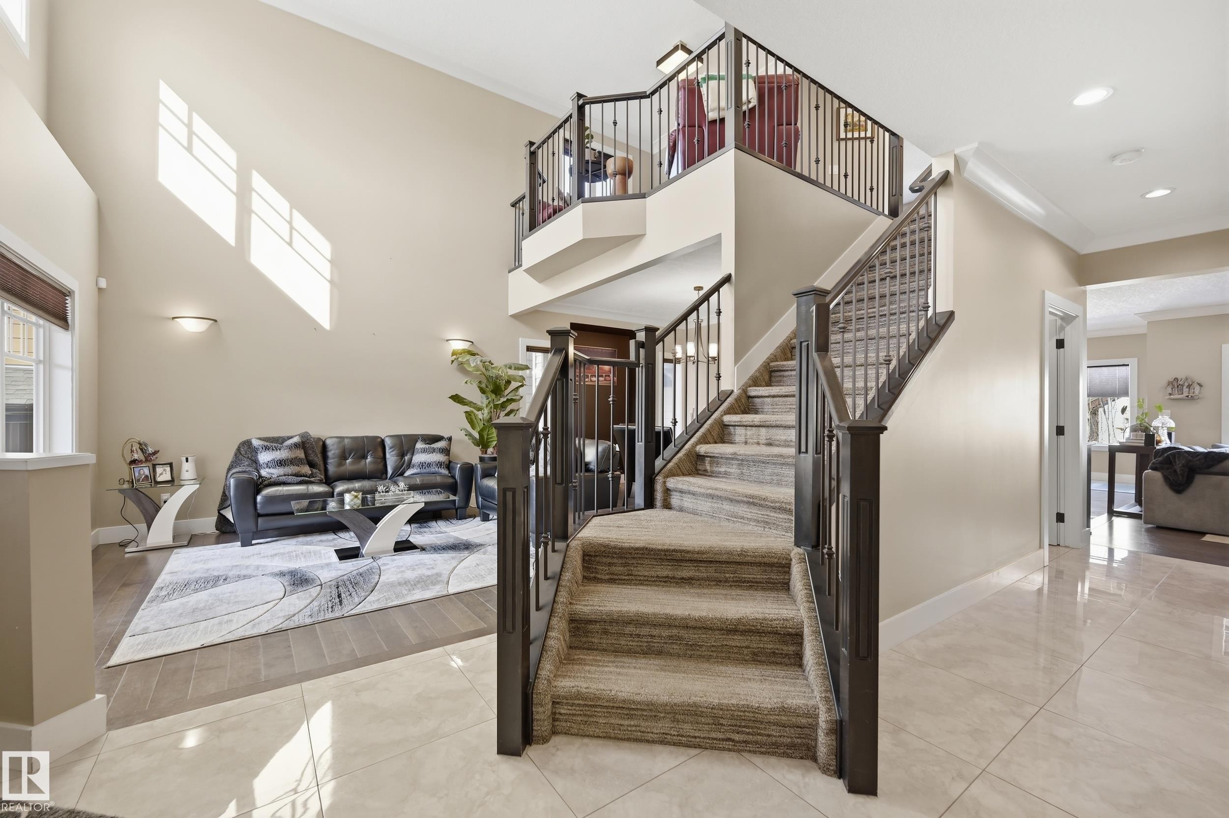 Expansive entryway featuring a grand staircase with carpeted treads and dark wood banisters, complemented by a two-story ceiling and natural light from high windows - 2018 Armitage Green, Edmonton, AB - Indoor Photo Showing Other Room