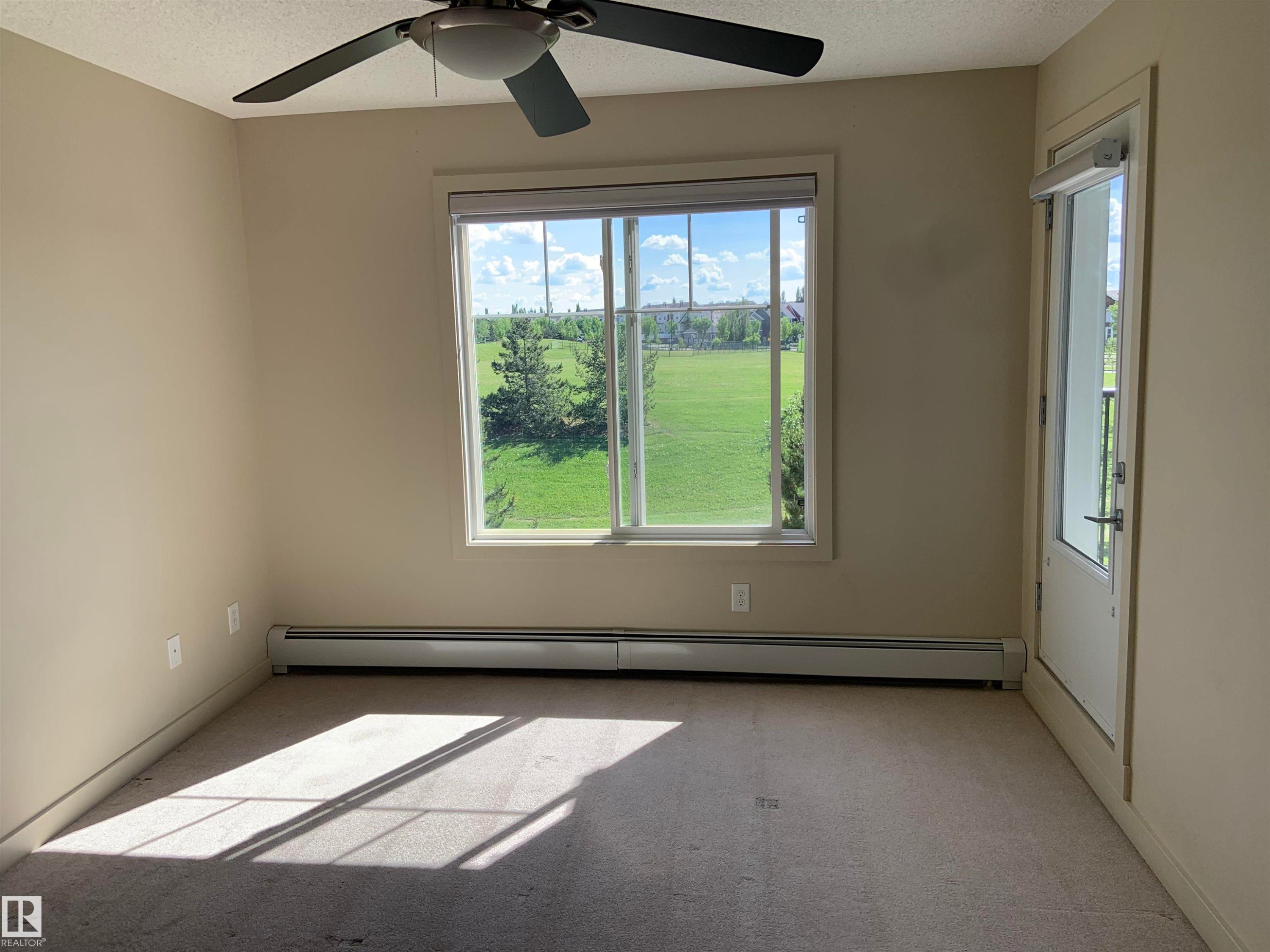 Interior room featuring neutral wall paint, carpet flooring, baseboard heating, a ceiling fan with integrated lighting, and a large window overlooking a green field - 307 6084 Stanton Drive, Edmonton, AB - Indoor Photo Showing Other Room