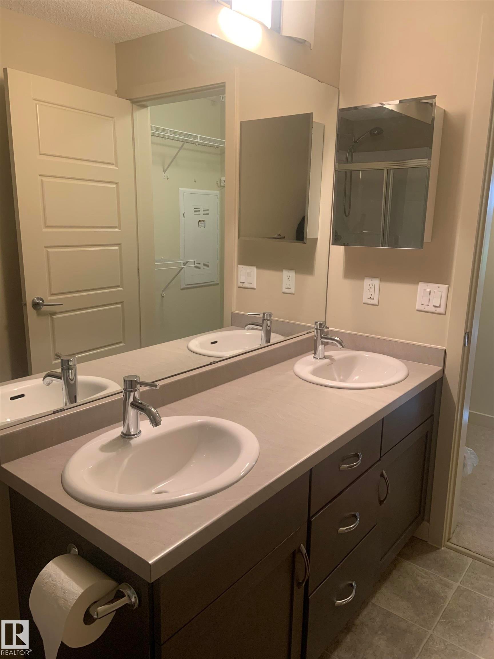 Bathroom featuring a dual-sink vanity with dark wood-finish cabinetry, light-toned countertops, and contemporary chrome faucets - 307 6084 Stanton Drive, Edmonton, AB - Indoor Photo Showing Bathroom