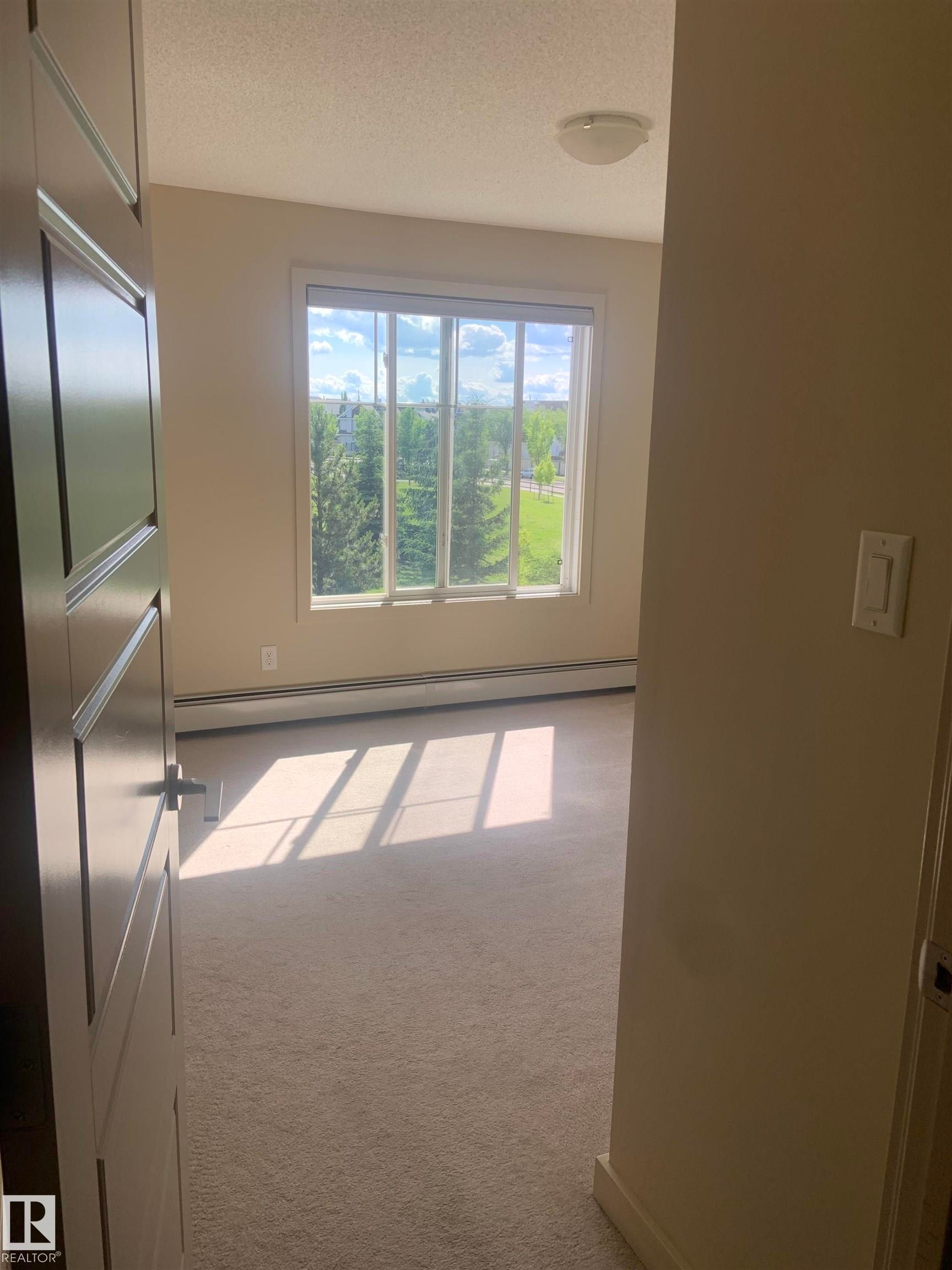 Spacious room featuring a large window, light neutral carpeting, baseboard heating, a contemporary flush-mount ceiling light fixture, and a solid-core paneled door - 307 6084 Stanton Drive, Edmonton, AB - Indoor Photo Showing Other Room