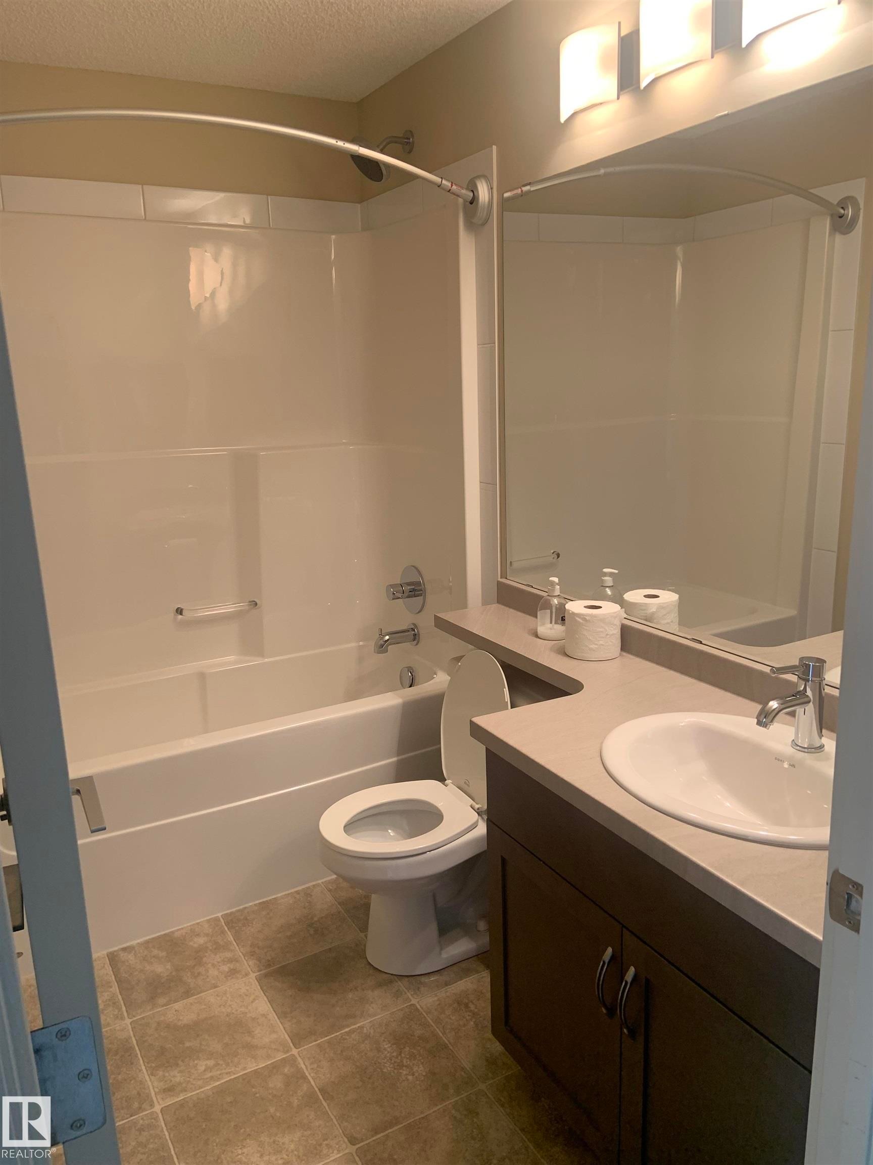 Bathroom featuring a white tub/shower combination with a curved shower rod, a dark wood-finish vanity with a built-in sink, a light-colored countertop, and a wall-mounted light fixture above a mirror - 307 6084 Stanton Drive, Edmonton, AB - Indoor Photo Showing Bathroom