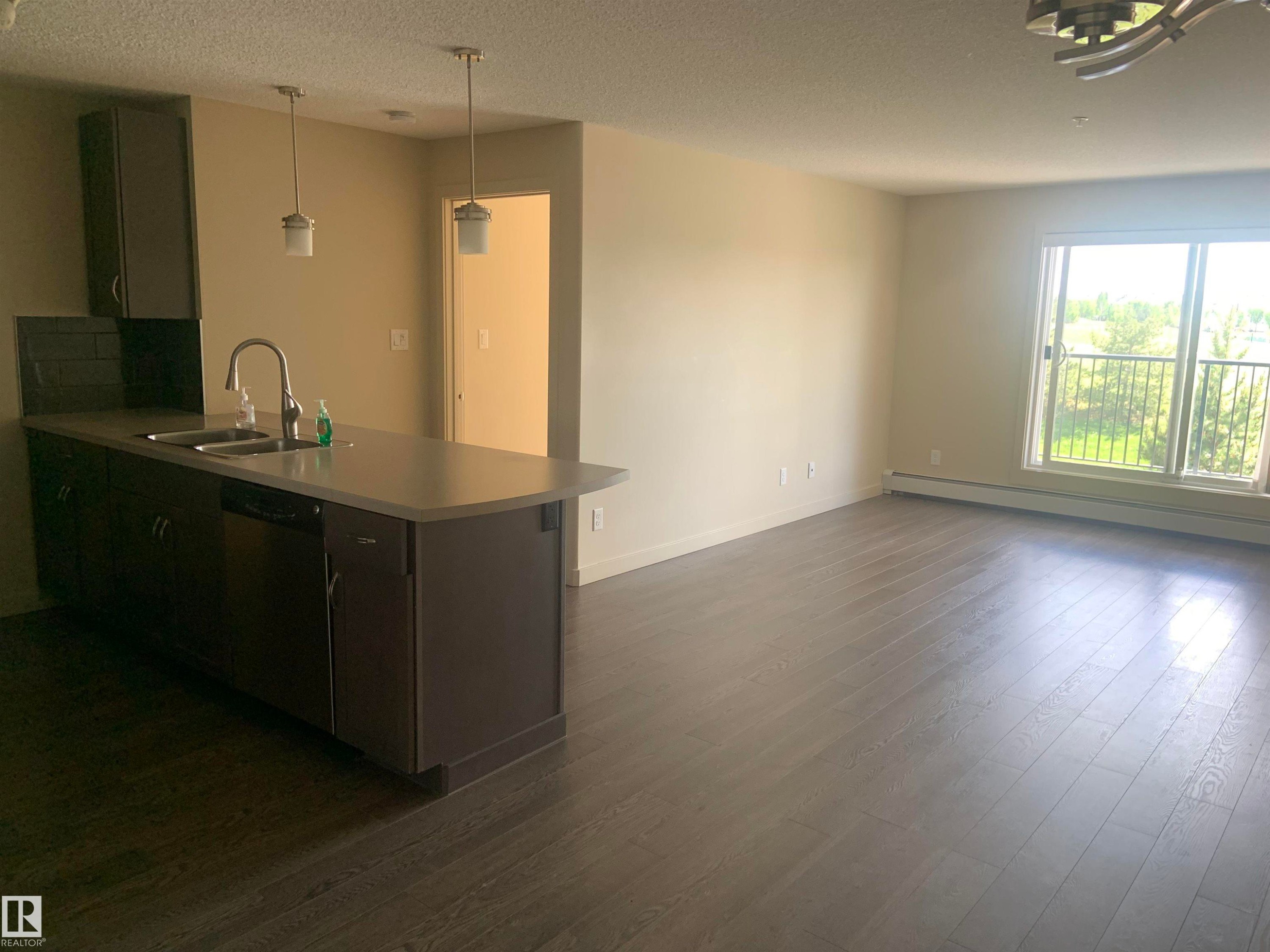 Open-concept living space featuring a kitchen island with integrated sink, dark wood-finish flooring, a sliding glass door to a balcony, and abundant natural light - 307 6084 Stanton Drive, Edmonton, AB - Indoor Photo Showing Kitchen With Double Sink