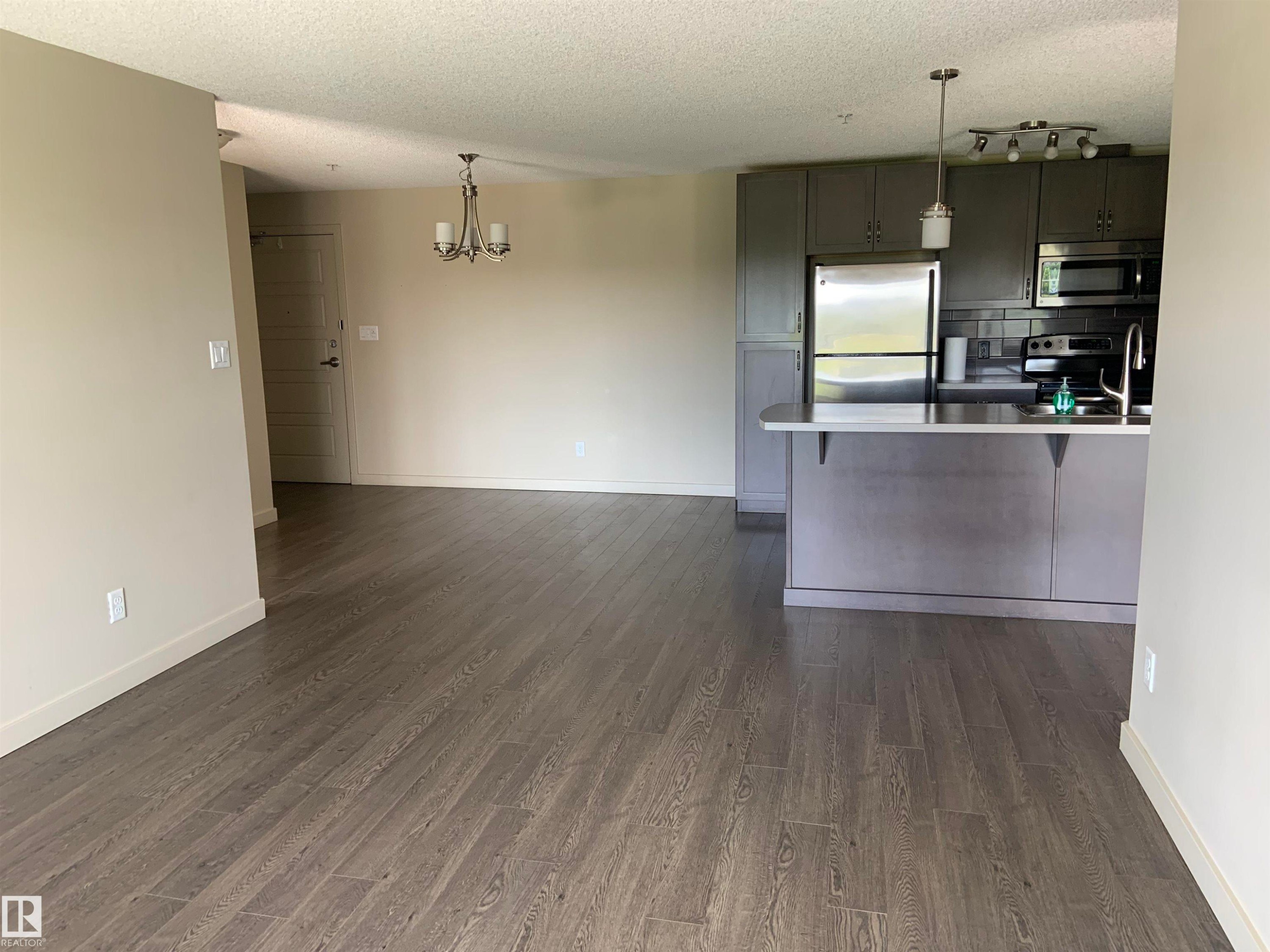 Open-concept living area featuring wood-finish flooring, a contemporary chandelier, and light neutral wall colors - 307 6084 Stanton Drive, Edmonton, AB - Indoor Photo Showing Kitchen