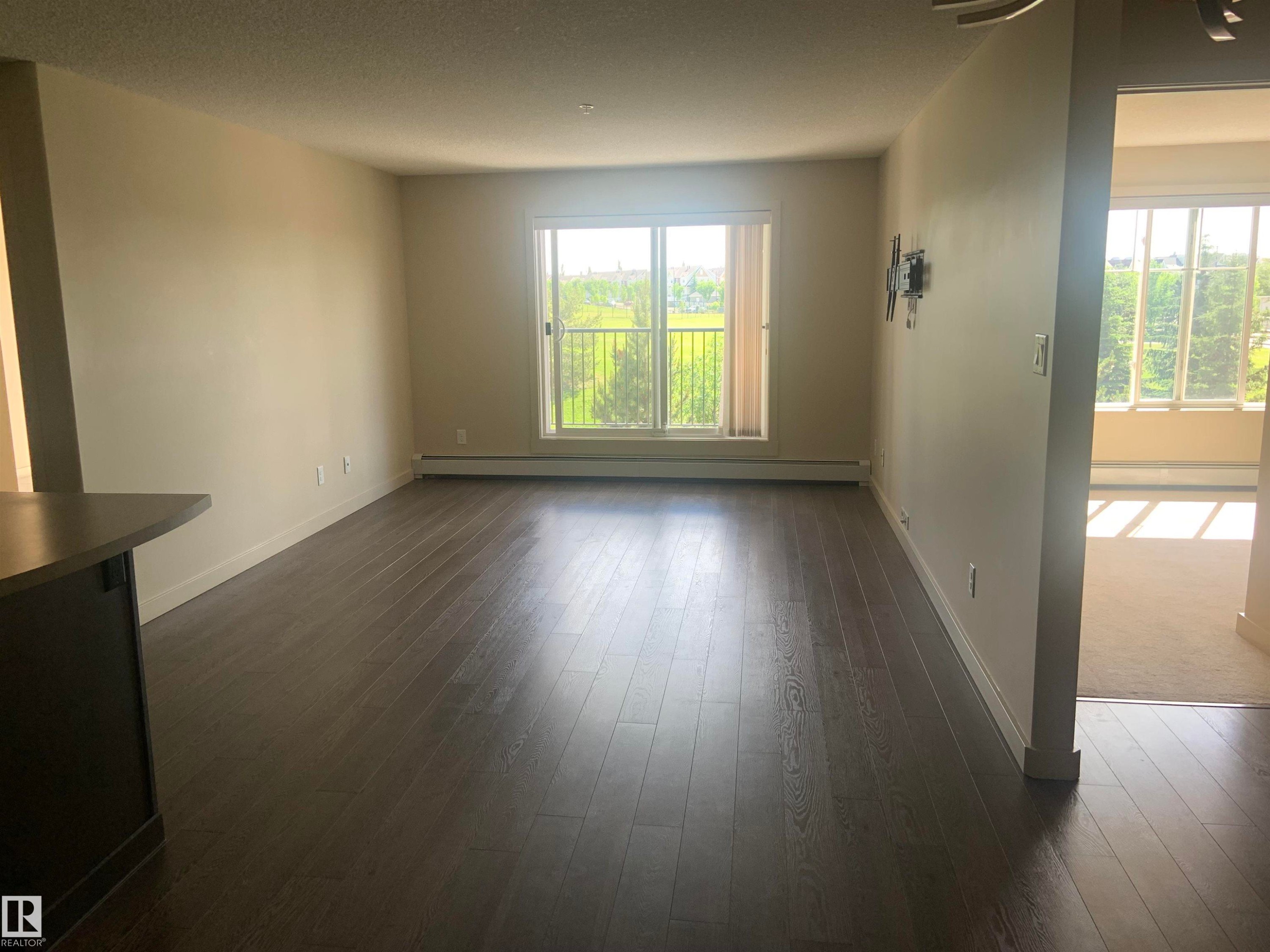 Spacious living area featuring dark wood-finish flooring, neutral wall paint, and a large sliding glass door leading to a balcony - 307 6084 Stanton Drive, Edmonton, AB - Indoor Photo Showing Other Room