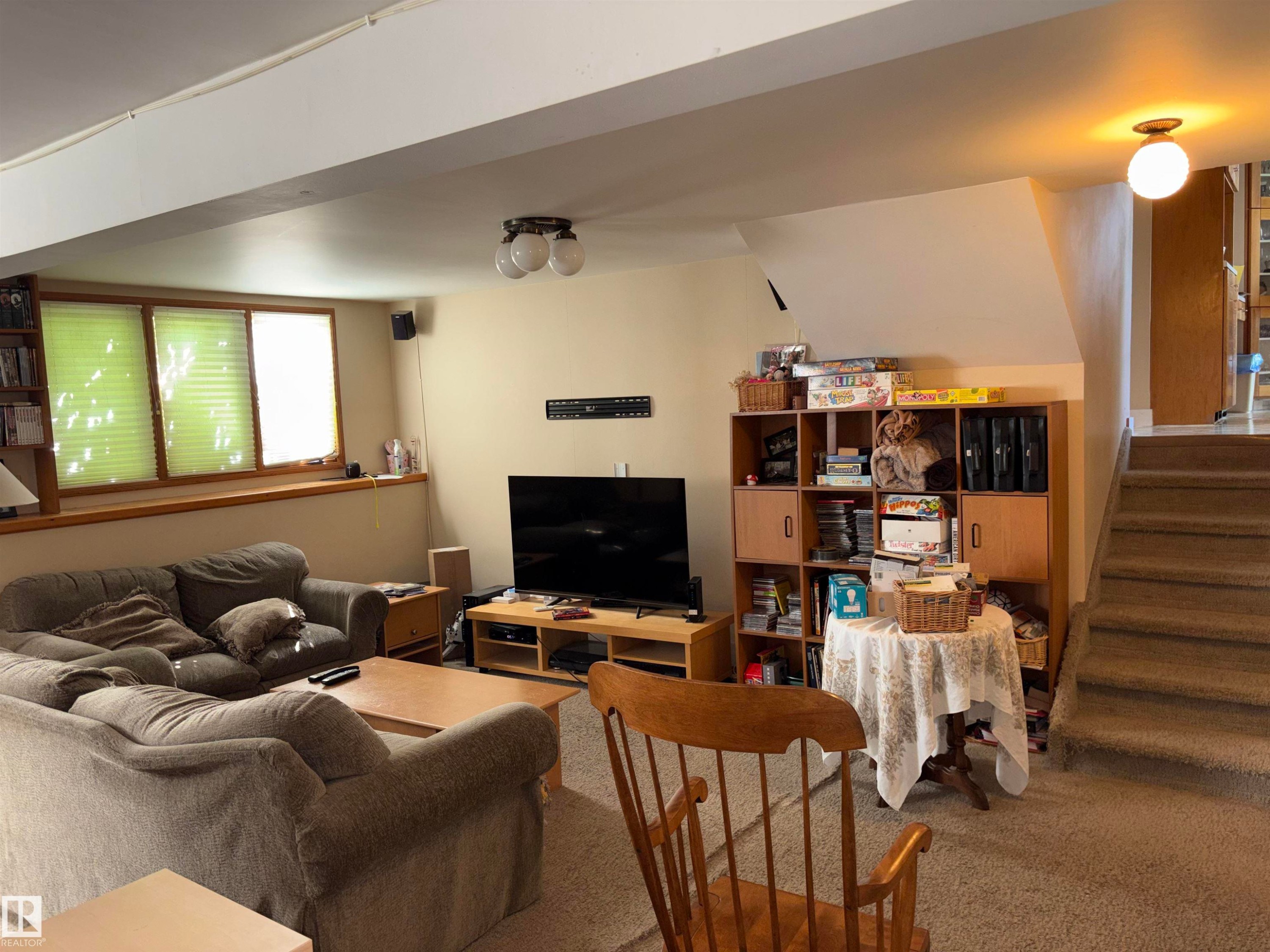 Carpeted recreation room featuring three windows with blinds, a built-in shelving unit, and a carpeted staircase - 8812 146 Street, Edmonton, AB