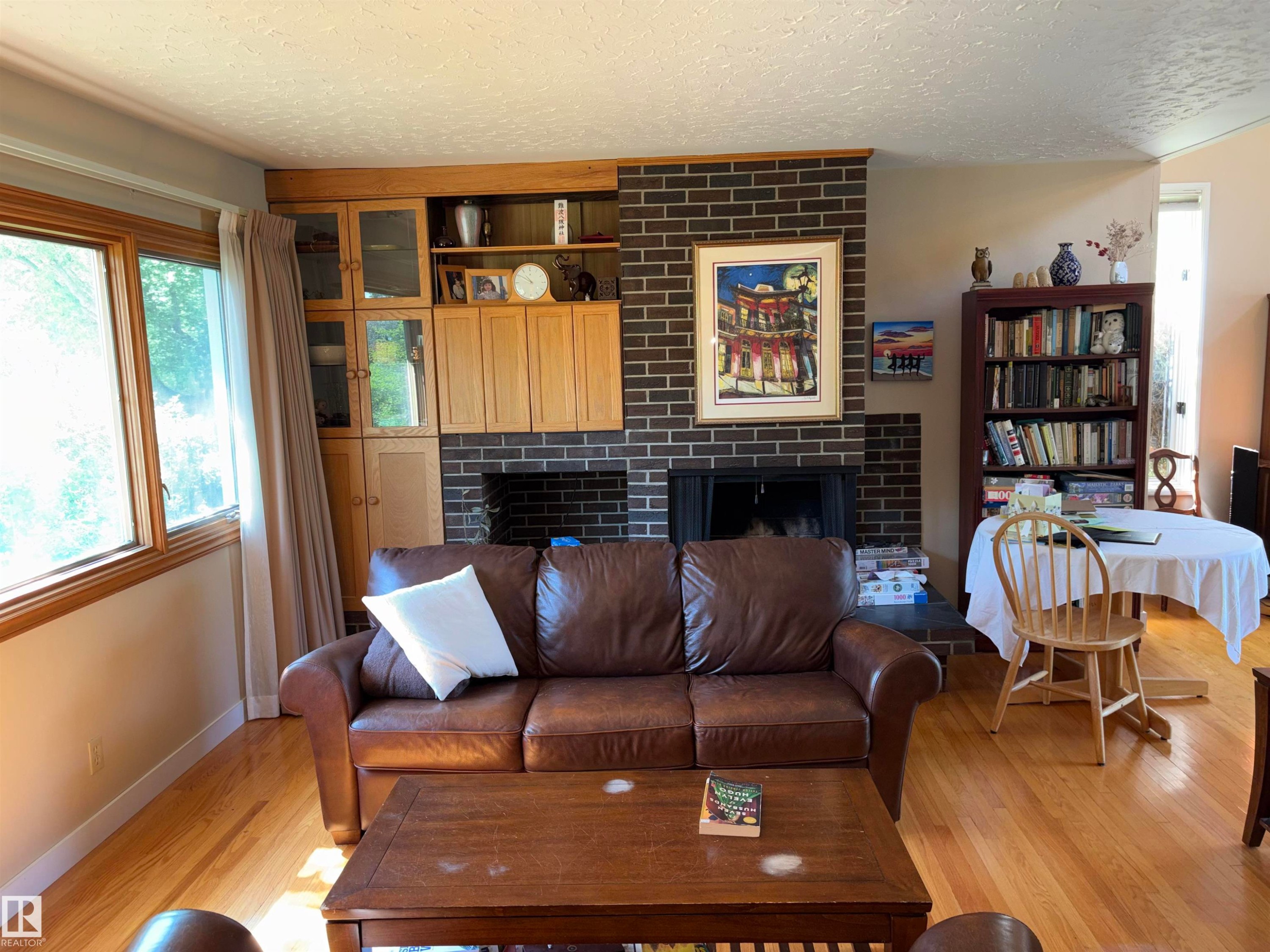 Living area featuring wood-finish flooring, a brick-surround fireplace, and a built-in cabinetry unit with glass-front and solid wood doors - 8812 146 Street, Edmonton, AB