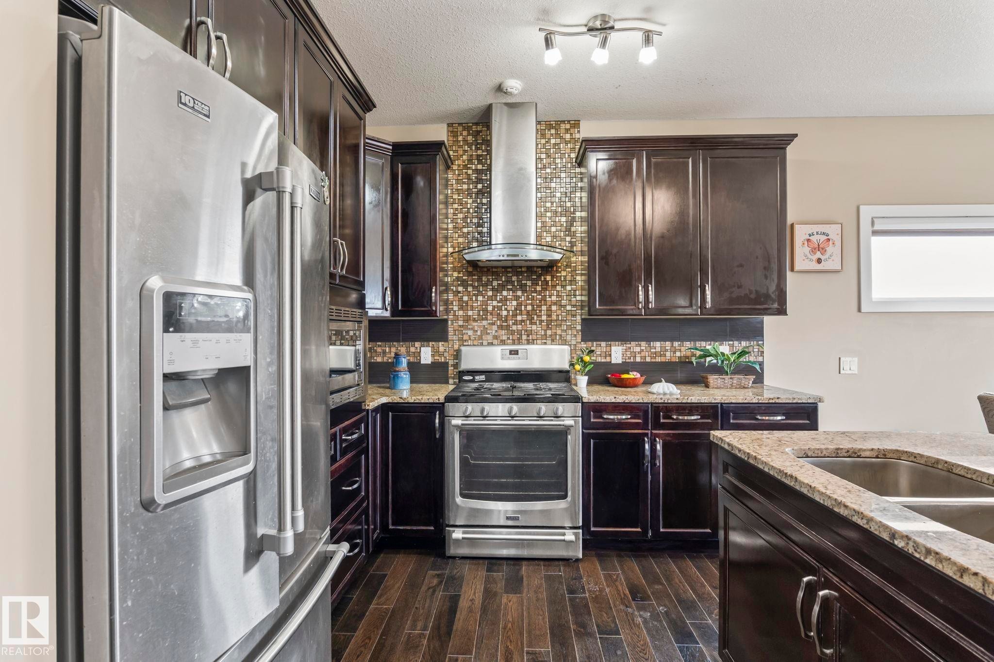 Kitchen featuring dark wood cabinetry, stainless steel appliances, a mosaic tile backsplash, wood-finish flooring, and granite countertops - 8431 216 St Nw Nw, Edmonton, AB - Indoor Photo Showing Kitchen