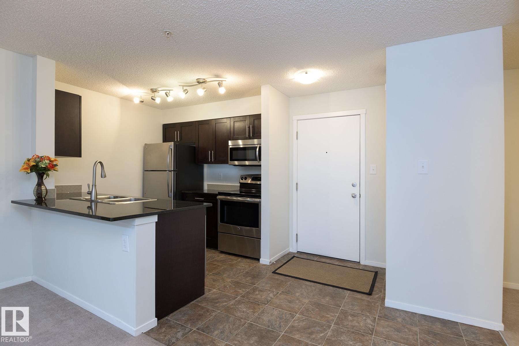 Kitchen and entryway featuring dark wood-finish cabinetry, stainless steel appliances, a breakfast bar with a dark countertop, and tiled flooring - 111 25 Element Dr, St. Albert, AB - Indoor Photo Showing Kitchen
