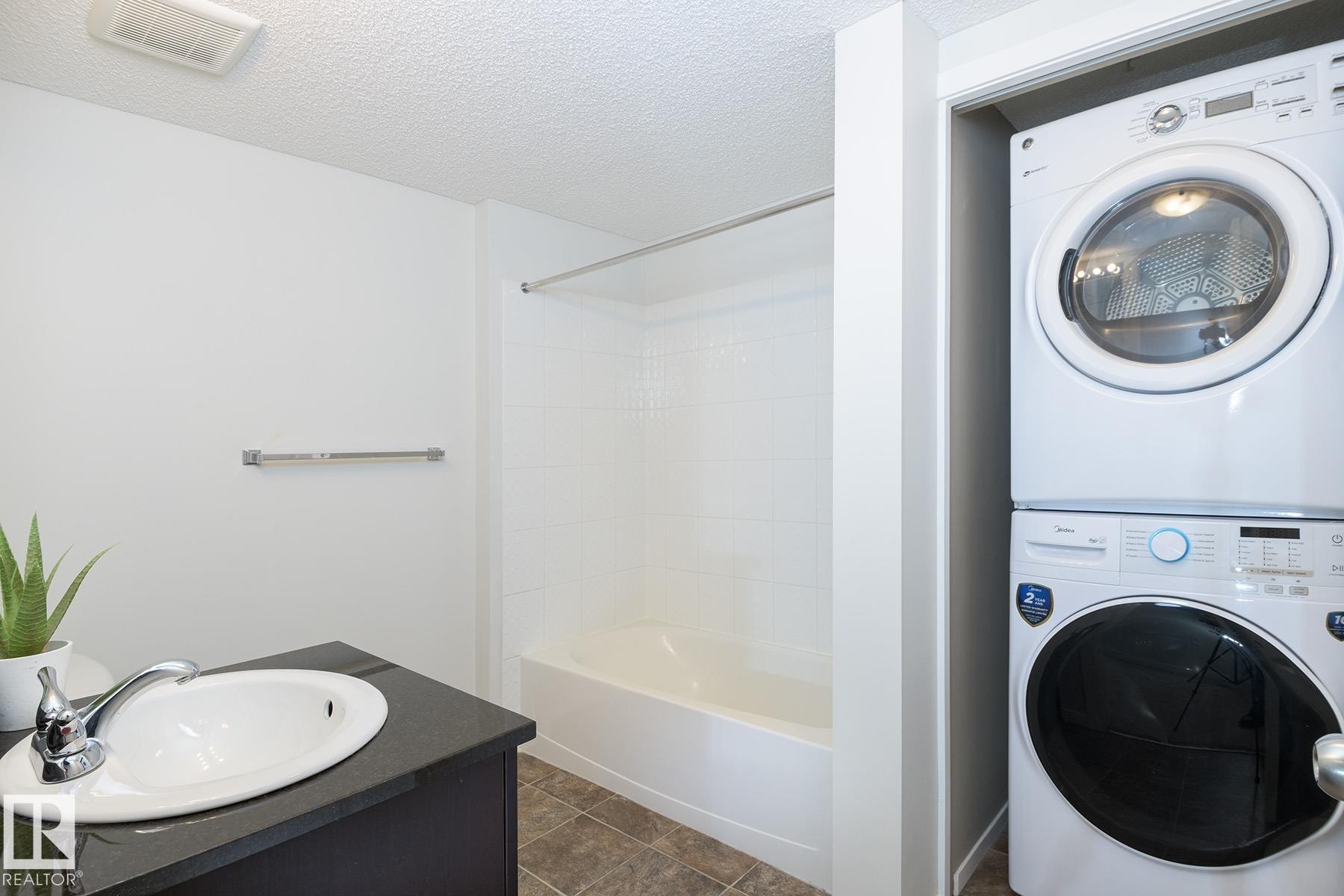 Bathroom featuring a built-in tub with tile surround, single vanity with oval sink, dark countertop, and stacked laundry appliances - 111 25 Element Dr, St. Albert, AB - Indoor Photo Showing Laundry Room