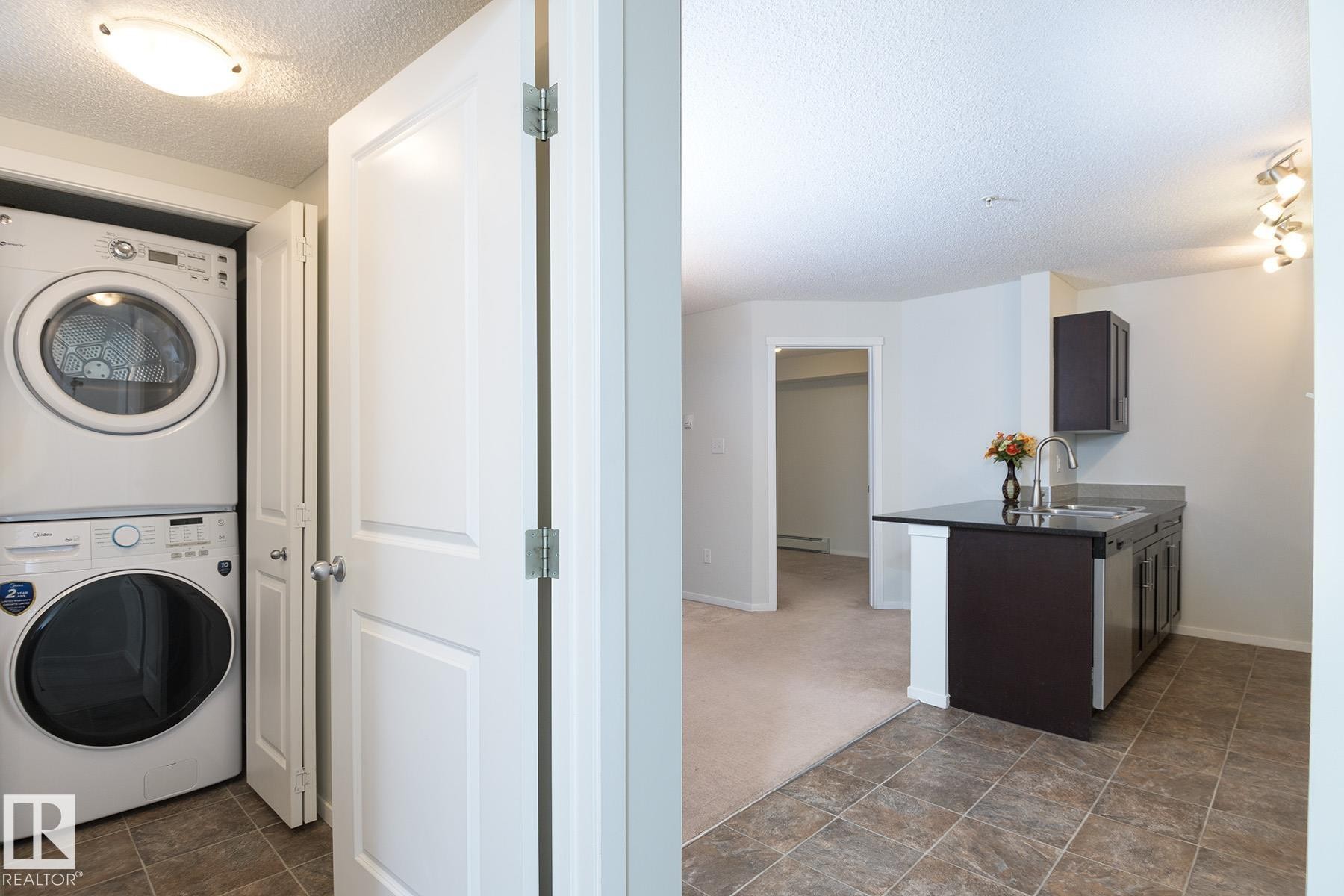 Compact laundry area featuring a stacked washer and dryer unit, complemented by a kitchen space with dark cabinetry, stainless steel sink, and a black countertop - 111 25 Element Dr, St. Albert, AB - Indoor Photo Showing Laundry Room